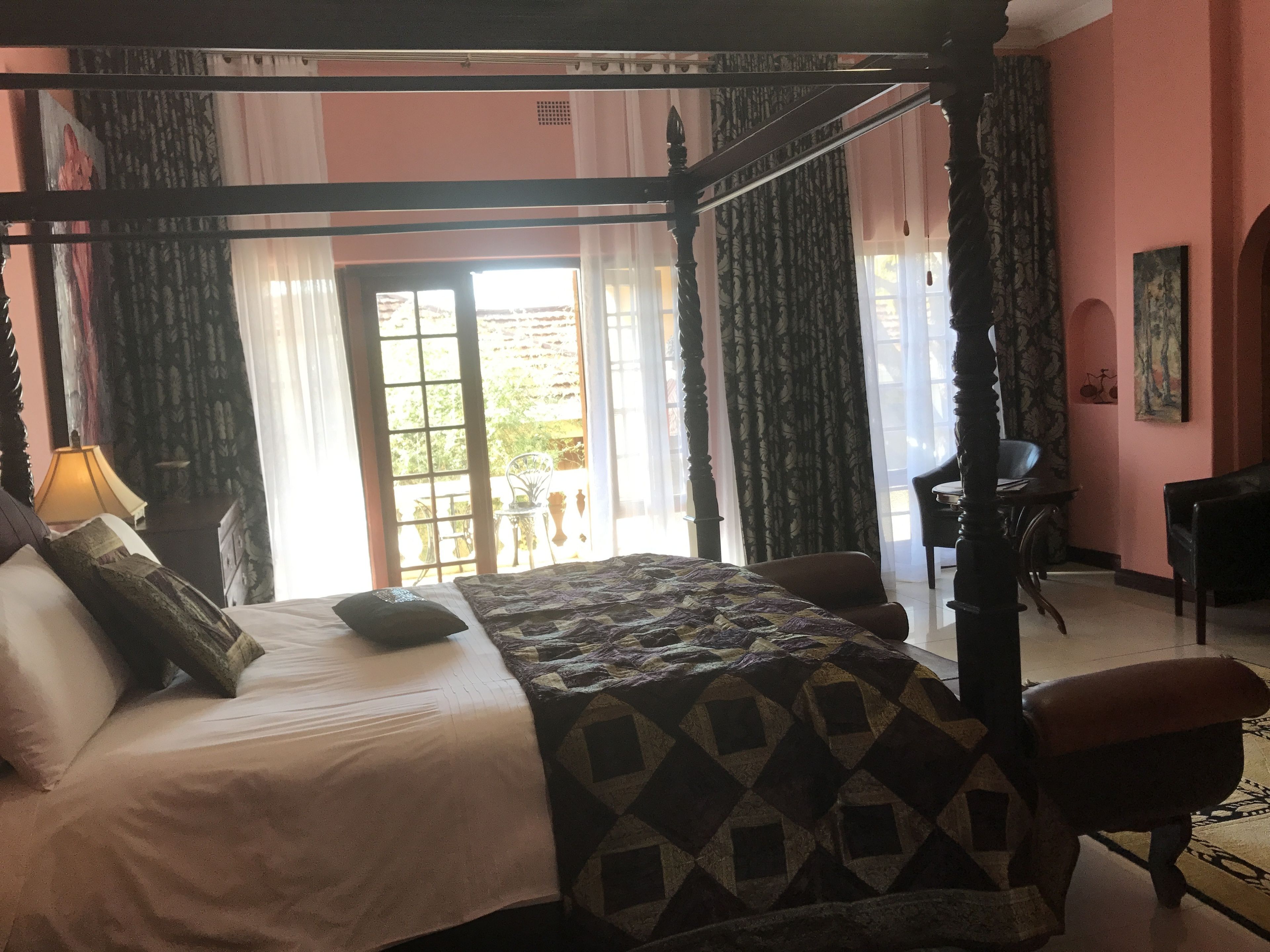 A four-poster bed with a patterned bedspread, a nightstand with a lamp, and a view of the outdoors through a large window.