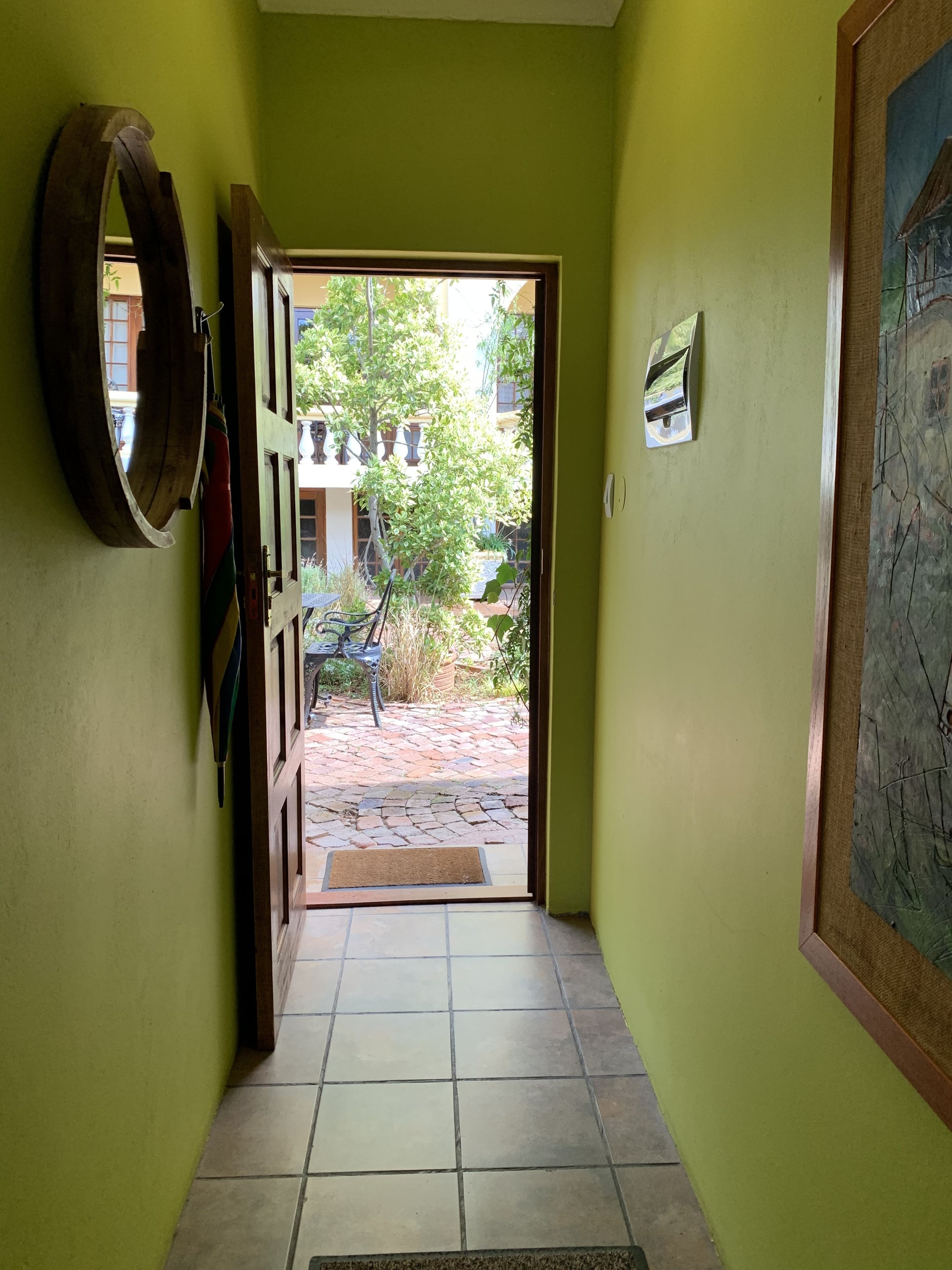 Un pasillo con pared verde, una puerta de madera que da a un patio con suelo de baldosas y un cuadro enmarcado a la derecha.