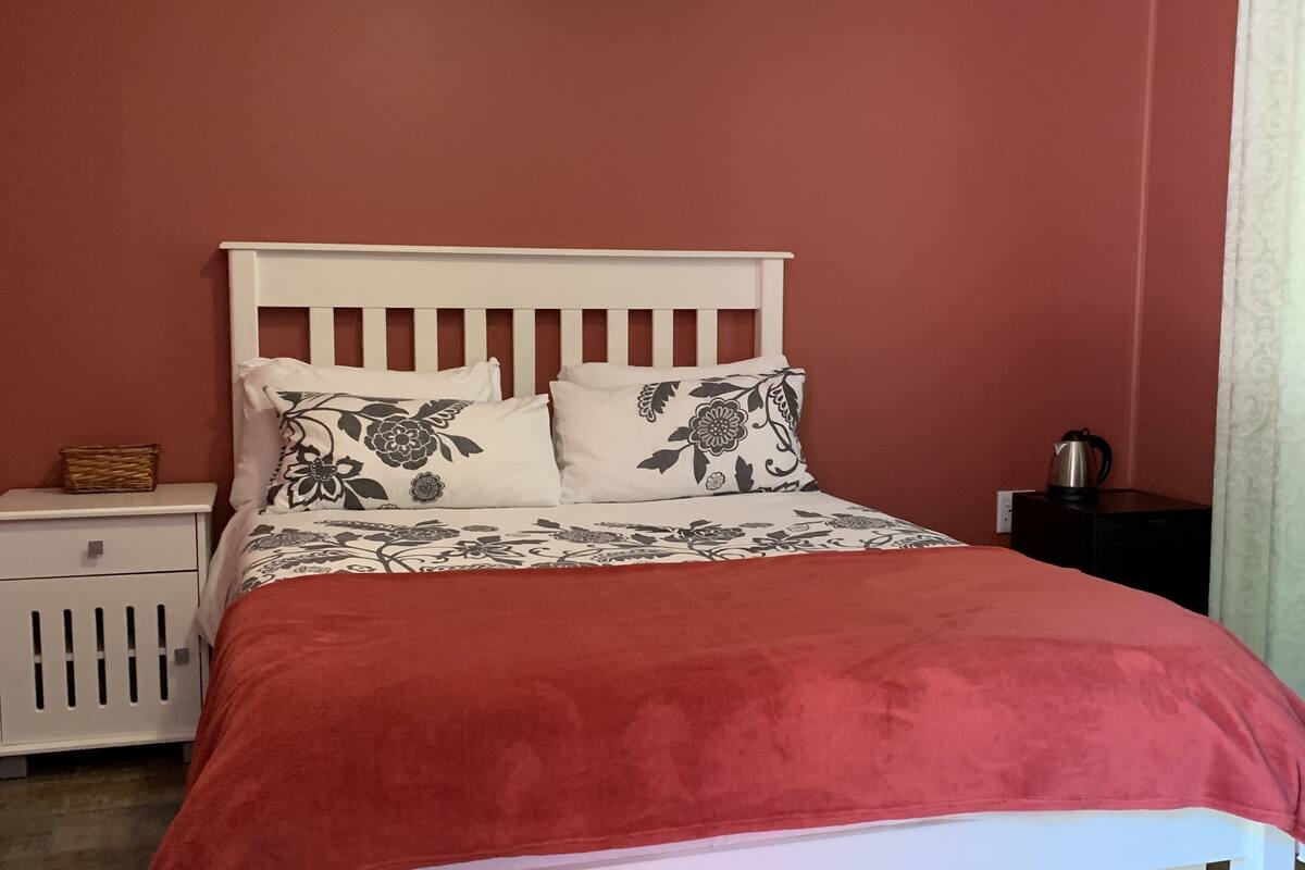 A bedroom with a white bed, red bedding, a ceiling fan, an air conditioning unit, a bedside table, and a nightstand.