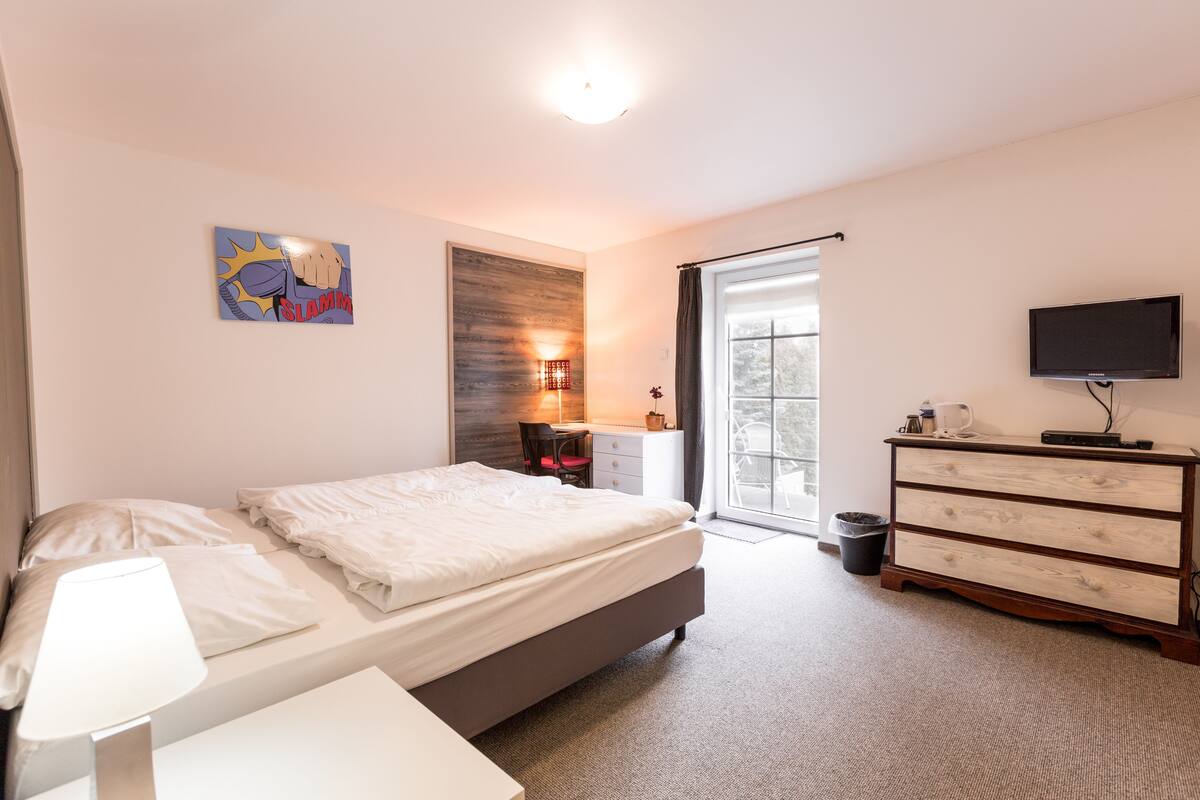 A bedroom with a bed, a desk, a chair, a dresser, a TV, and a window.