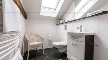 A modern bathroom with a skylight, white tiles, a wicker chair, a toilet, and a vanity with a dark cabinet.