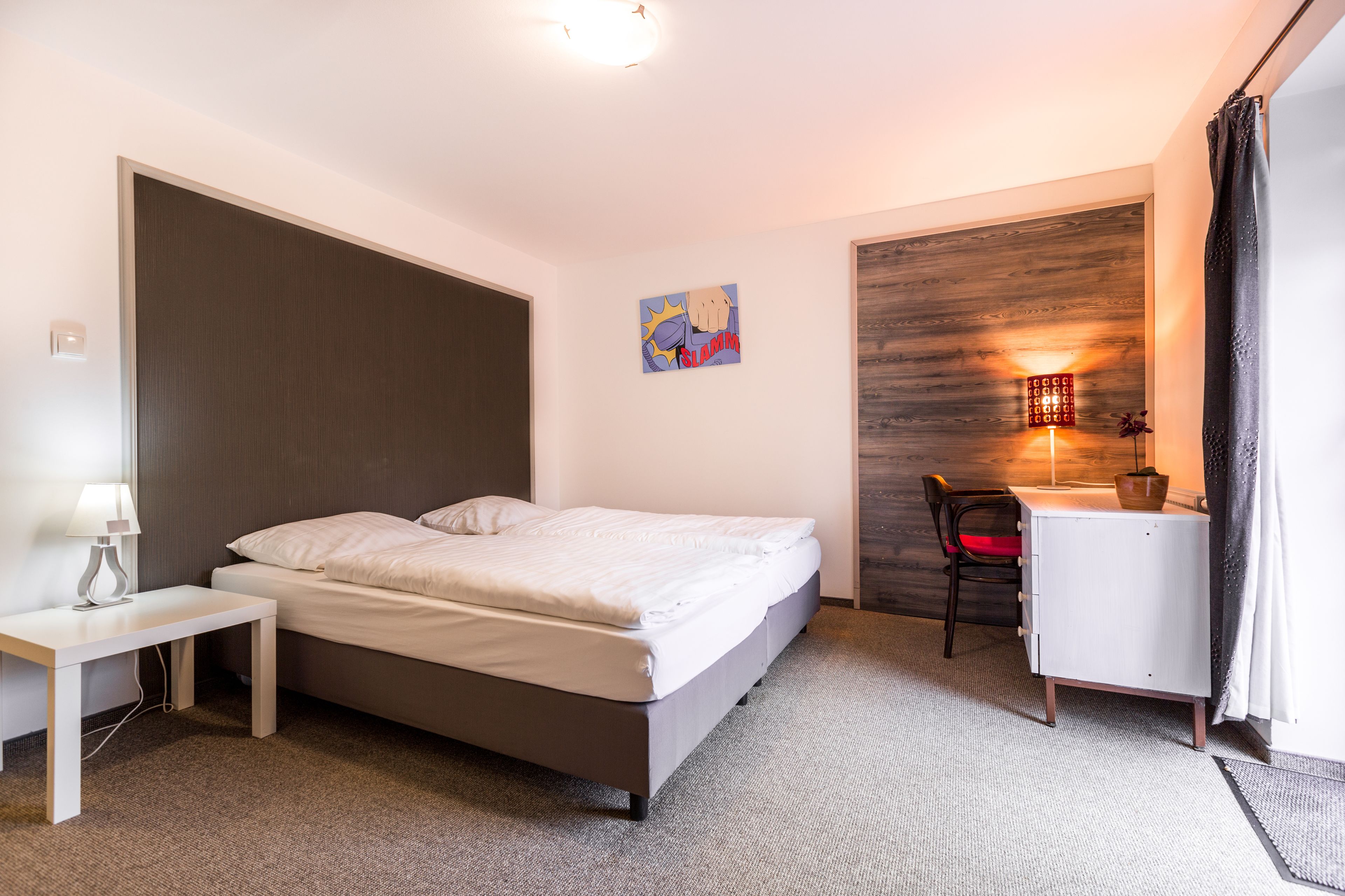 Hotellihuone, jossa on sänky, yöpöytä, työpöytä, tuoli ja seinään kiinnitetty valaisin.