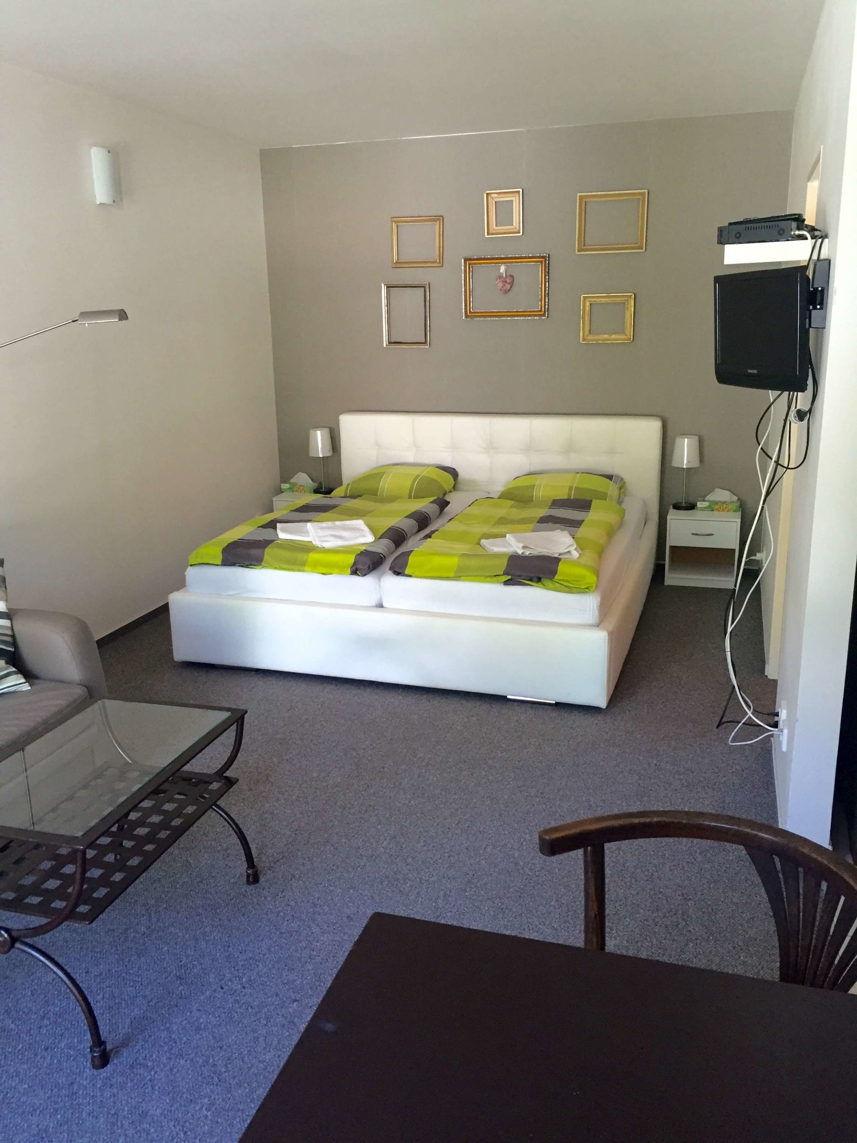 A bedroom with a bed, a glass table, a chair, a TV, and framed pictures on the wall.