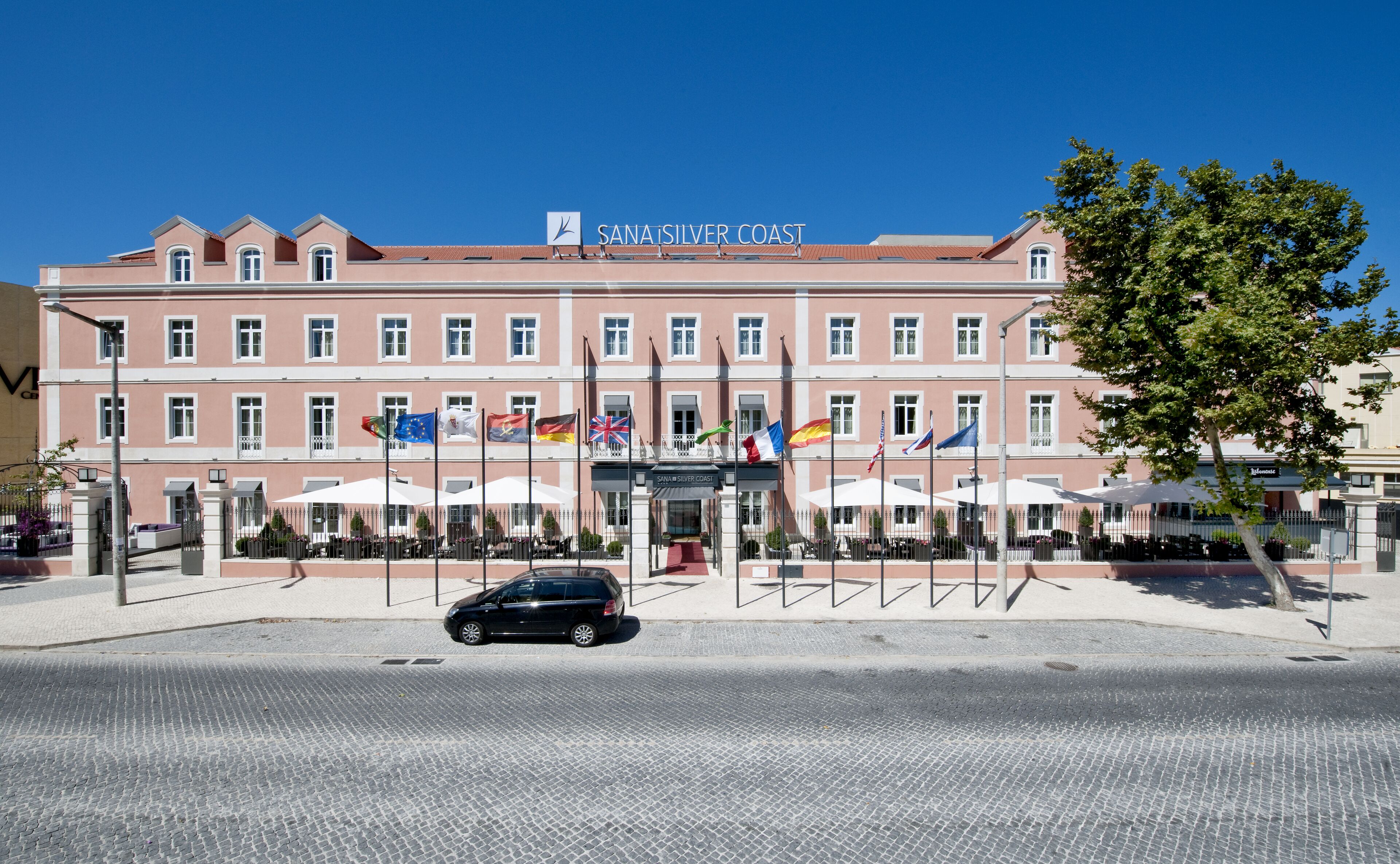 Un hotel con fachada rosa, varias banderas y mesas al aire libre.
