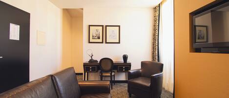 A hotel room with a leather sofa, a desk with a chair, a television, and framed pictures on the wall.
