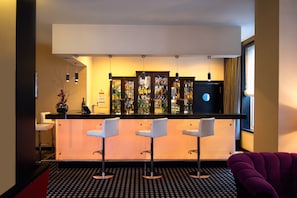 A modern bar area with a bar counter, three white bar stools, and a variety of bottles and glasses.