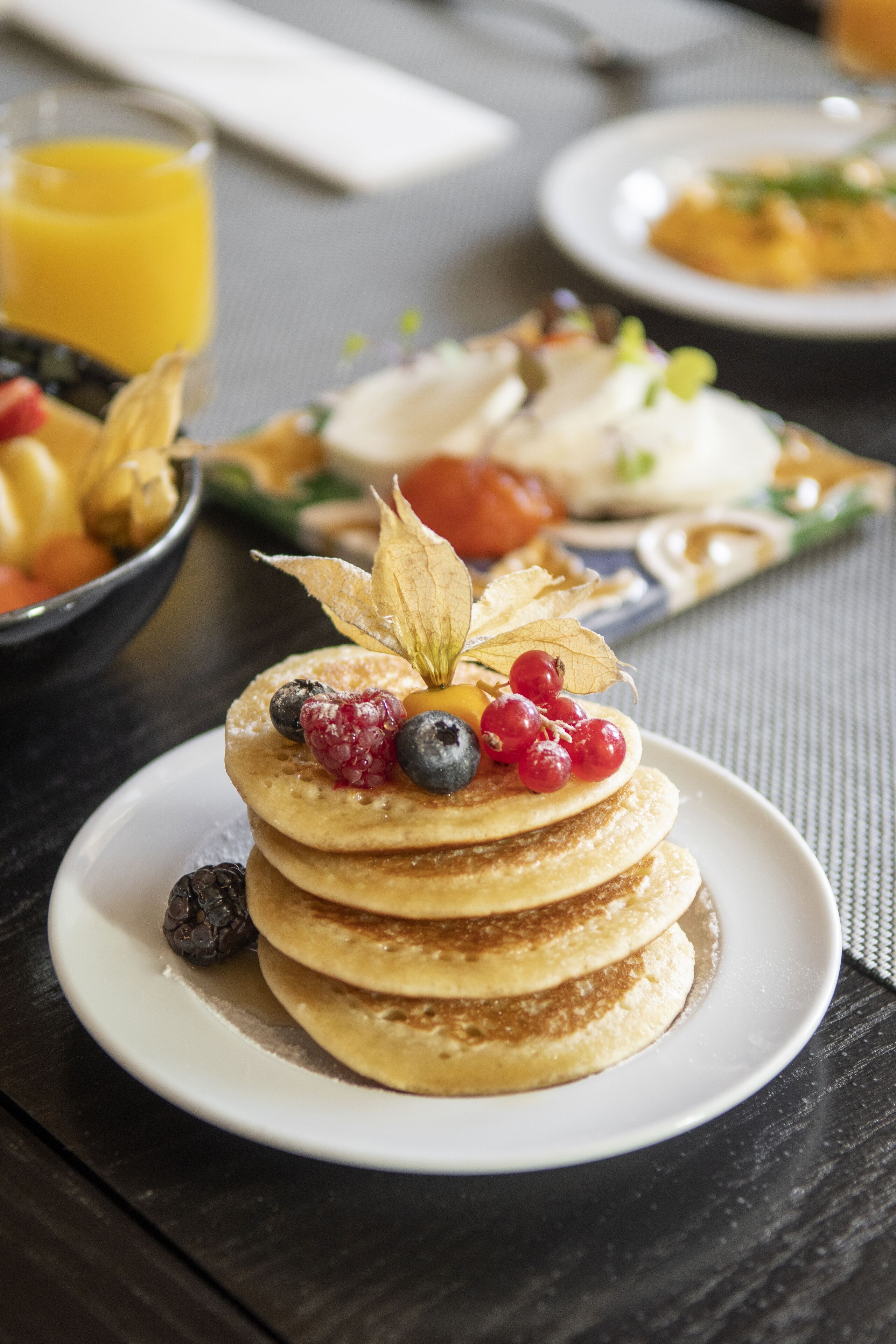 Desayuno con tortitas cubiertas de frutos rojos y una hoja, un vaso de zumo de naranja y un bol de fruta sobre una mesa.