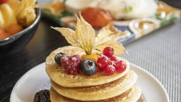A breakfast setting with pancakes topped with berries and a leaf, a glass of orange juice, and a bowl of fruit on a table.