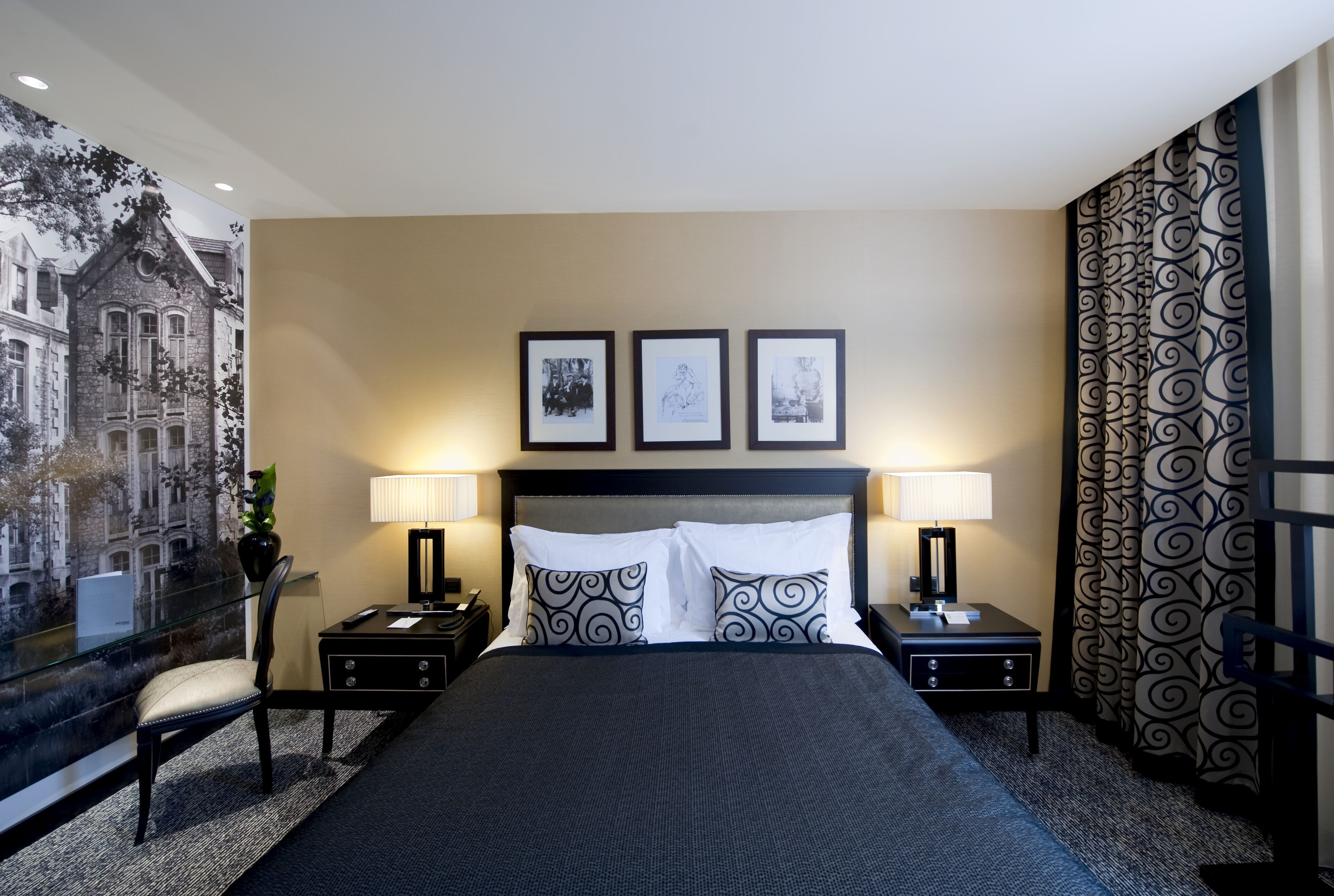 Habitación de hotel con una cama grande, dos mesitas de noche con lámparas, una silla, un ventanal con vistas a un edificio y tres cuadros enmarcados en la pared.