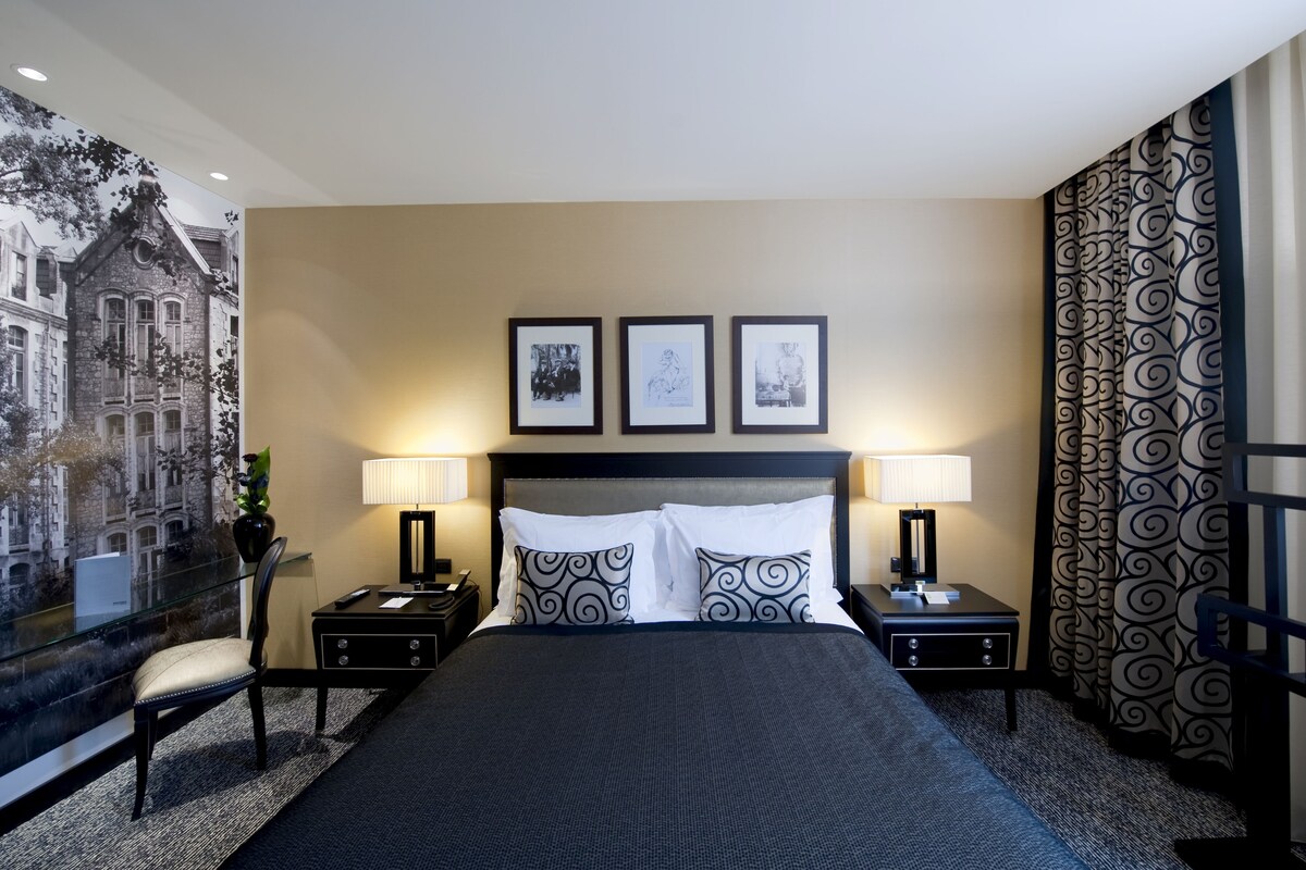 A hotel room with a large bed, two bedside tables with lamps, a chair, a large window with a view of a building, and three framed pictures on the wall.