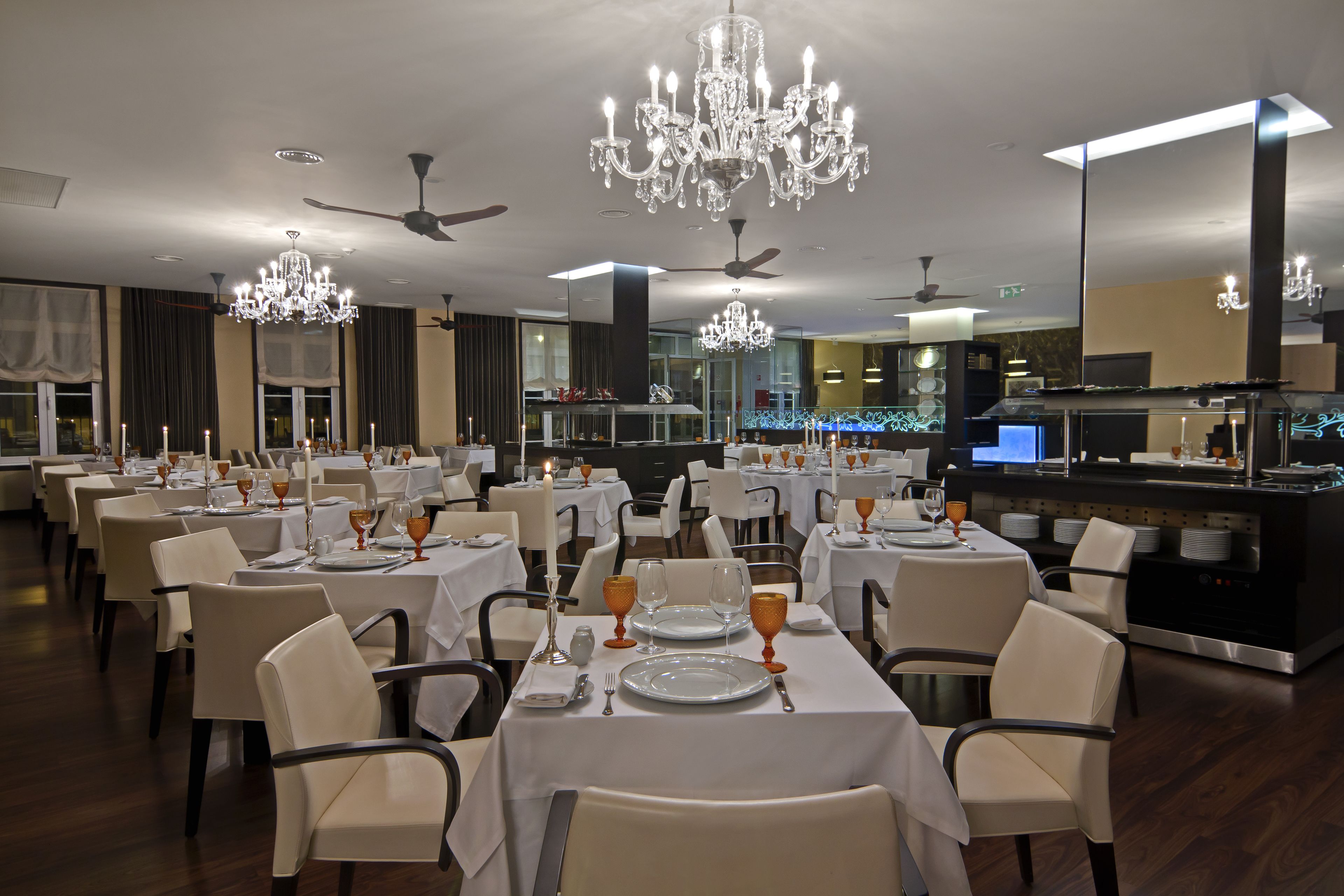 Un restaurante con varias mesas dispuestas para comer, candelabros, ventiladores de techo y una zona de cocina al fondo.