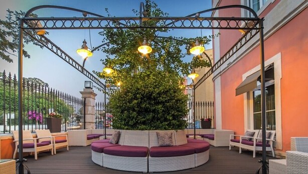 Et cirkulært udendørs siddeområde med et træ i midten, omgivet af fletningsmøbler og en metalpergola med lys.