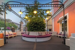 A circular outdoor seating area with a central tree, surrounded by wicker furniture and a metal pergola with lights.