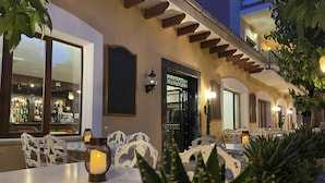 An outdoor seating area with white ornate chairs and tables, a lit lantern, and a building entrance with a sign. - Hostal Villa Rosa (Calvia)