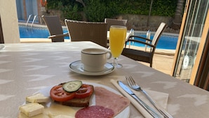 A breakfast setting with a plate of cheese, ham, and salami, a glass of orange juice, and a cup of coffee on a table by a pool. - Hostal Villa Rosa (Calvia)