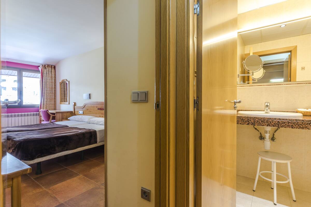 A hotel room with two beds, a bathroom with a sink and mirror, and a window with curtains.