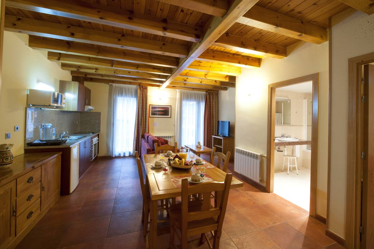 A spacious kitchen with wooden beams, a dining area with a table set for four, and a view into a bathroom.