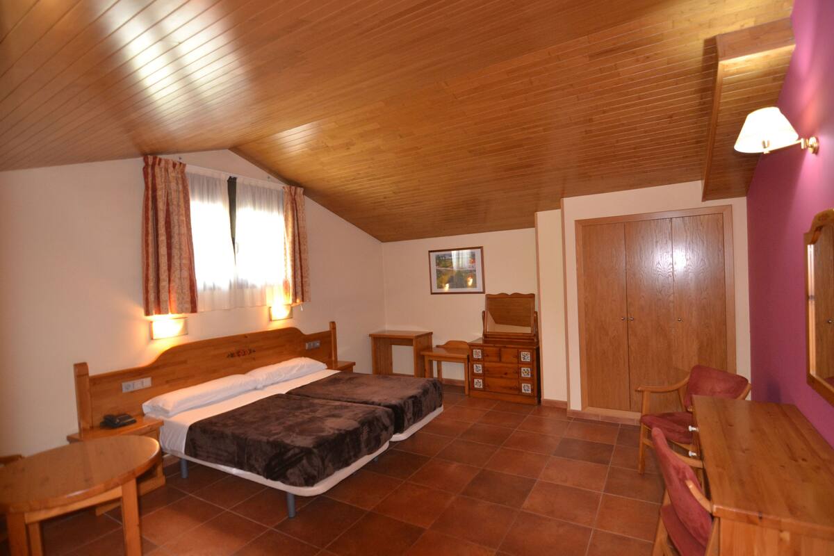A bedroom with two beds, a wooden ceiling, a wooden wardrobe, a small table, and a chair.