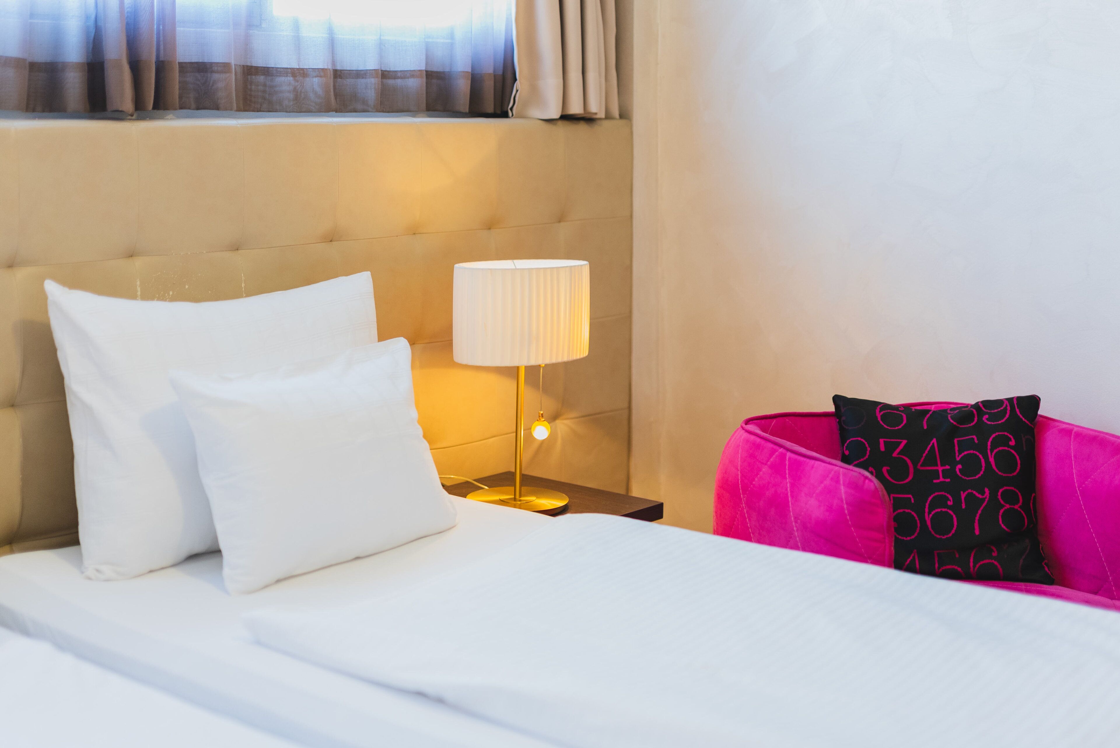 A hotel room with a bed, a nightstand with a lamp, a pink chair, and a window with curtains.