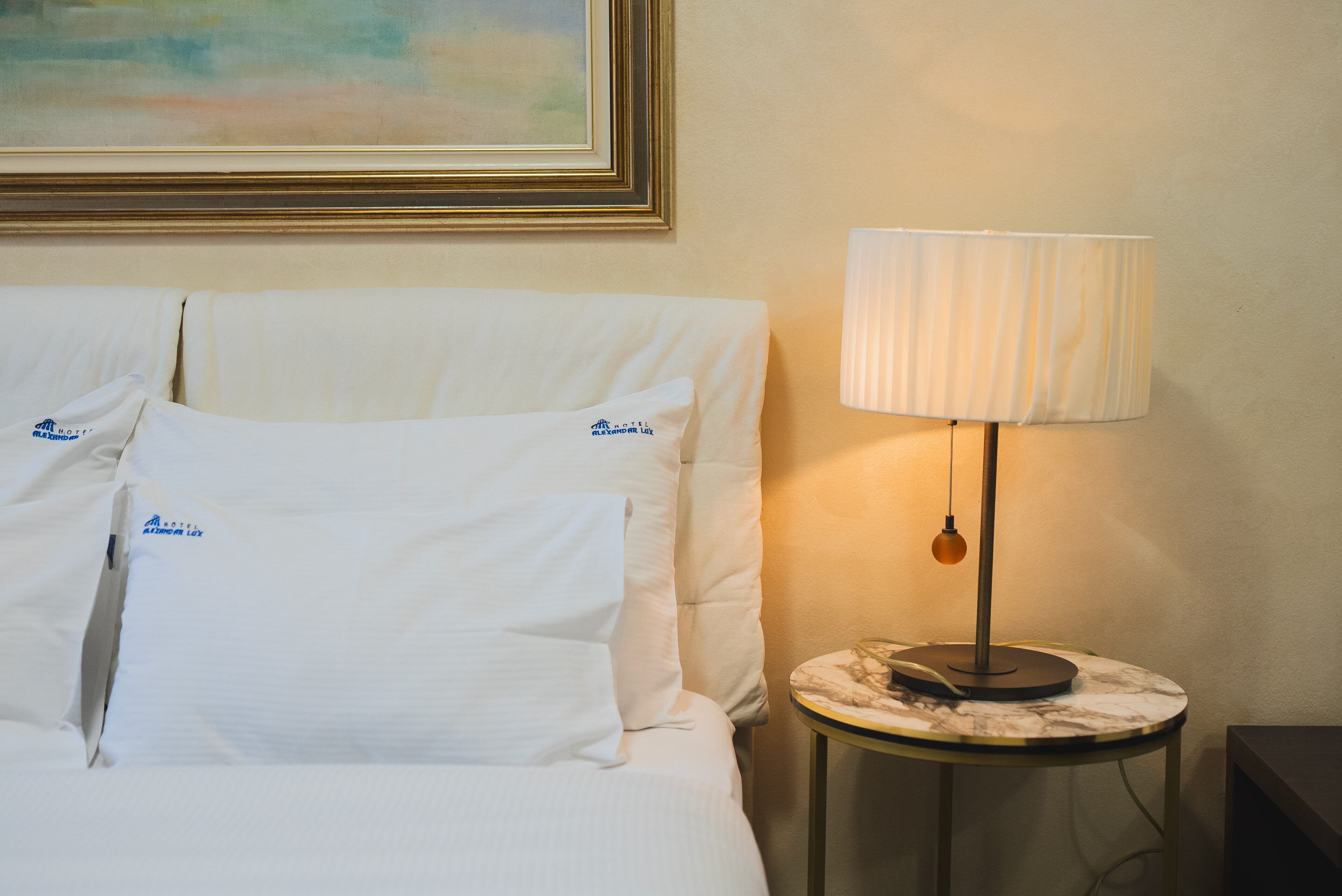 A neatly made bed with white linens and pillows, a round side table with a lamp, and a framed painting on the wall.