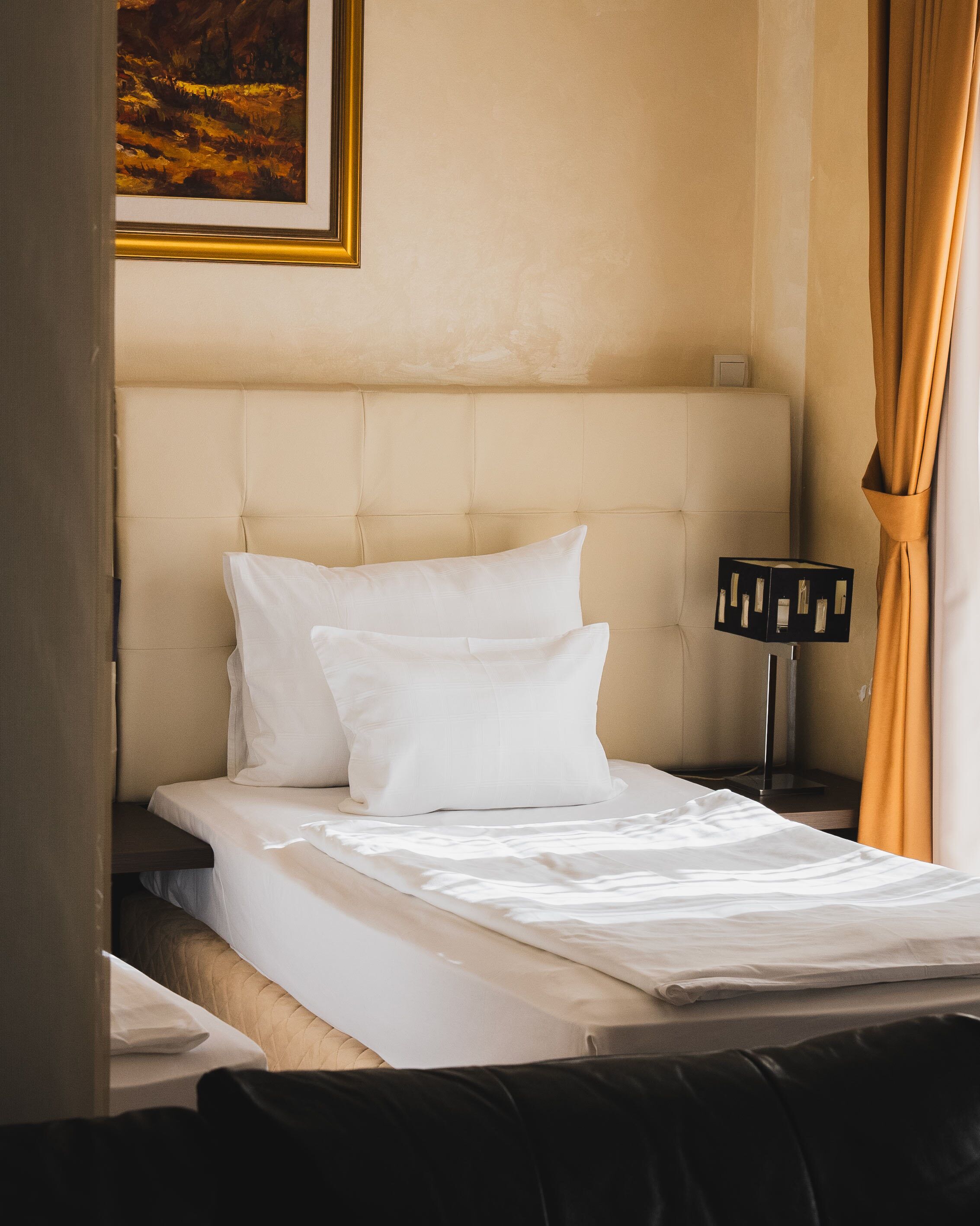 Una cama bien hecha con sábanas y almohadas blancas, un cabecero, una lámpara de noche y un cuadro en la pared.