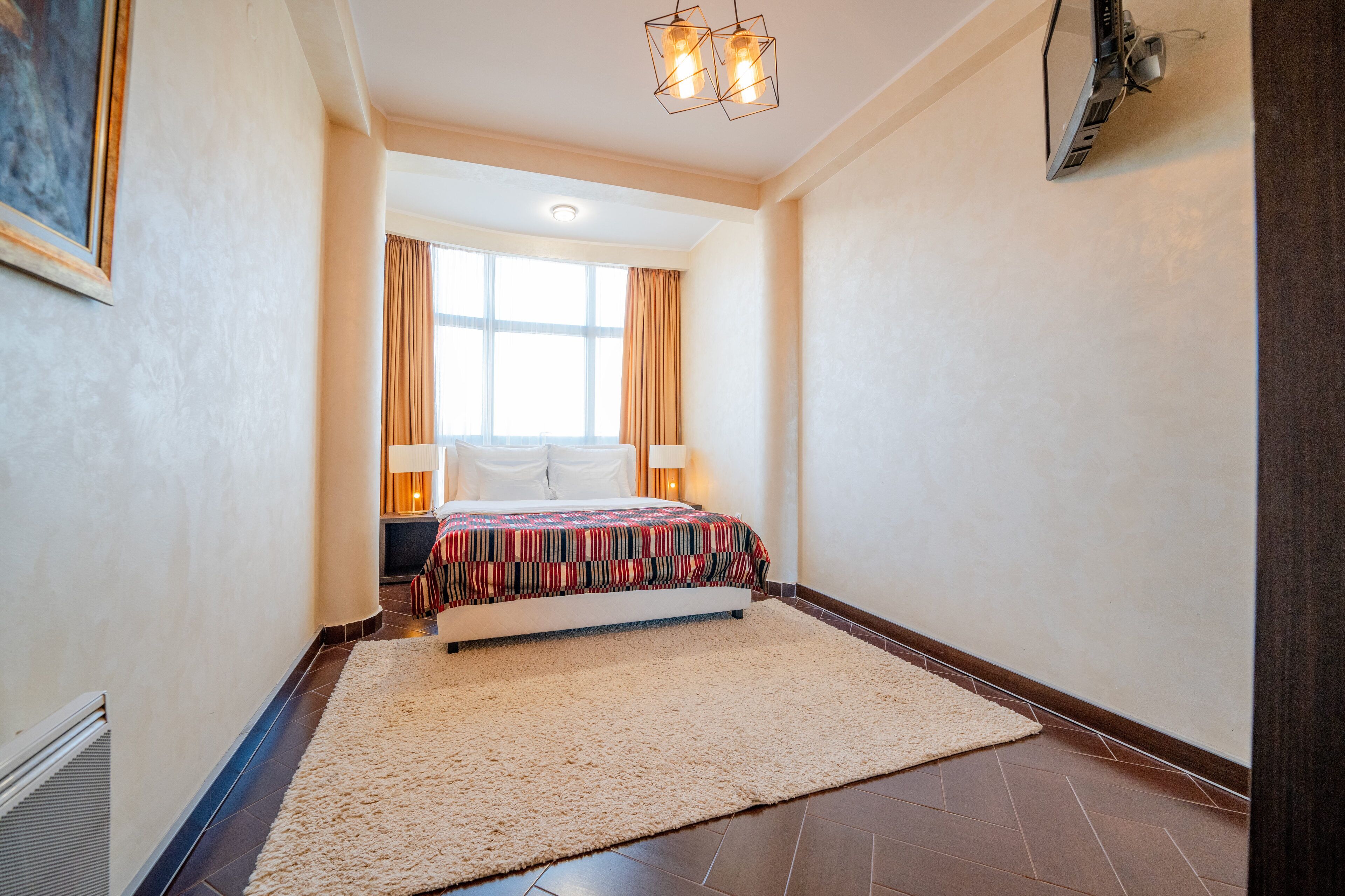 A hotel room with a bed, a large window, a nightstand, and a wall-mounted TV.