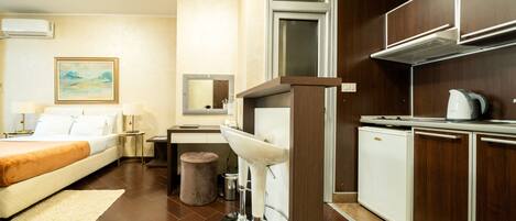 Una habitación de hotel con cama, mesita de noche, lámpara y una kitchenette con lavamanos y un refrigerador pequeño.