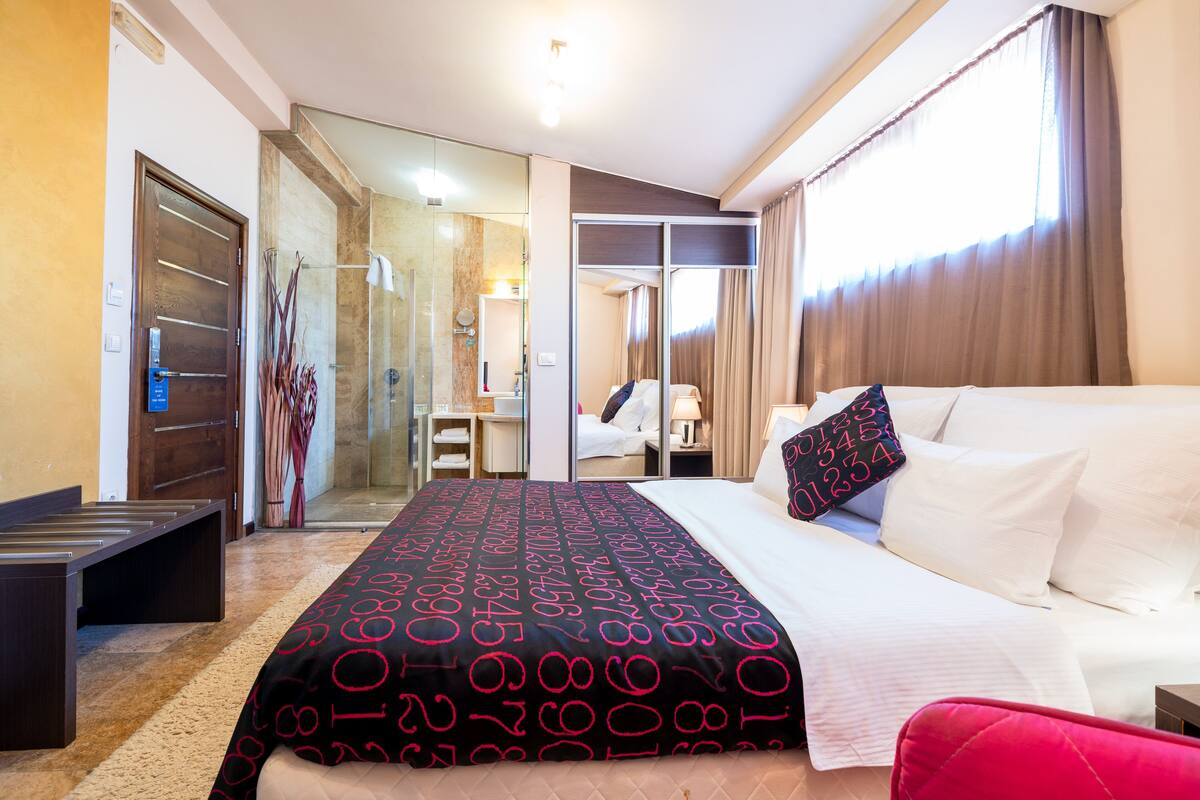 A hotel room with a bed, a desk, a chair, and a bathroom visible through a glass door.