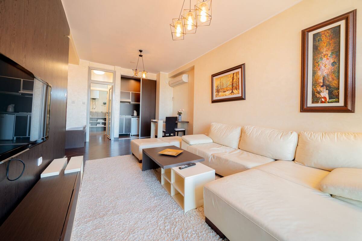 A modern living room with a sofa, coffee table, and TV. There is a kitchen area in the background.
