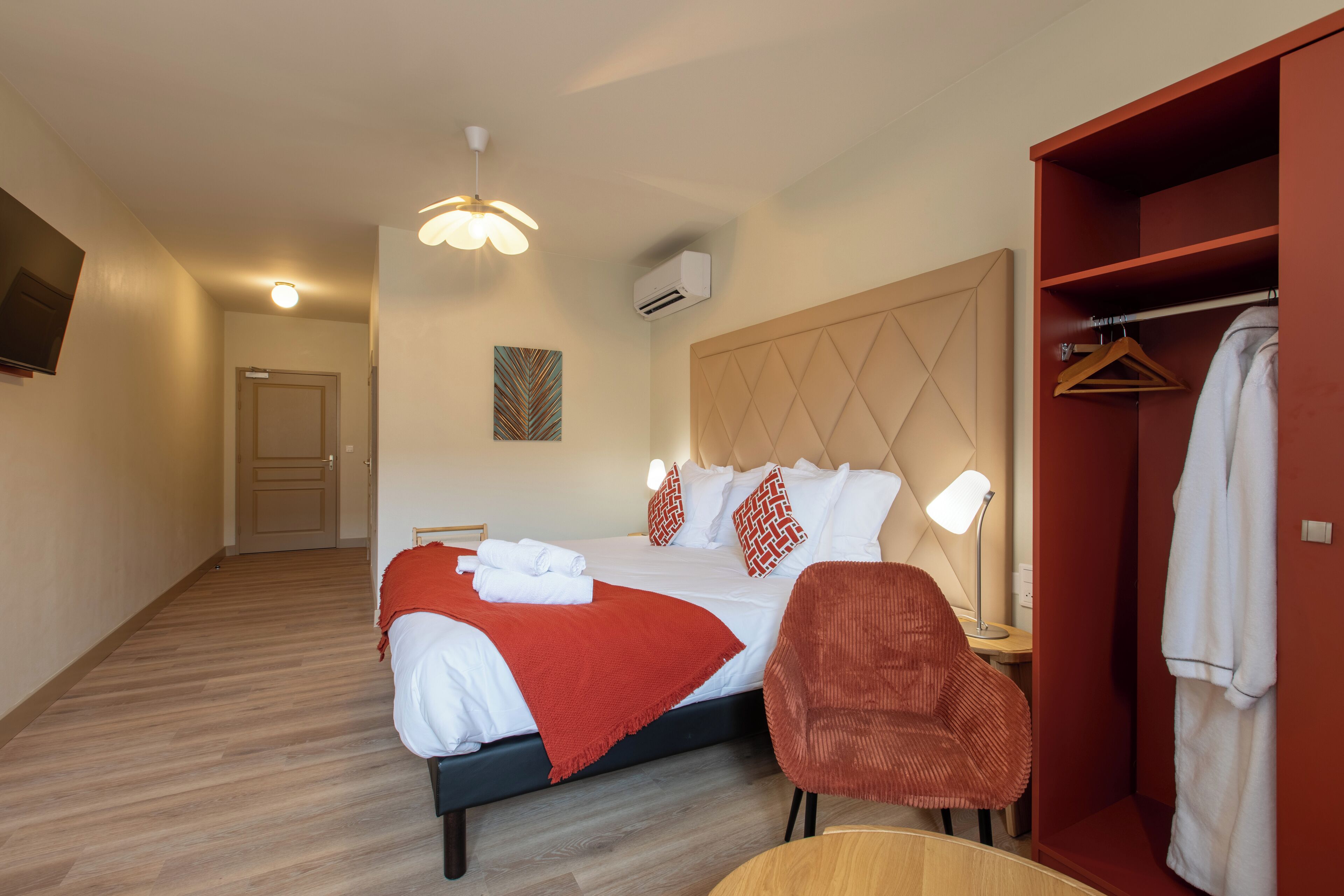 A hotel room with a bed, a red chair, a wooden side table, a robe, and a coat rack.
