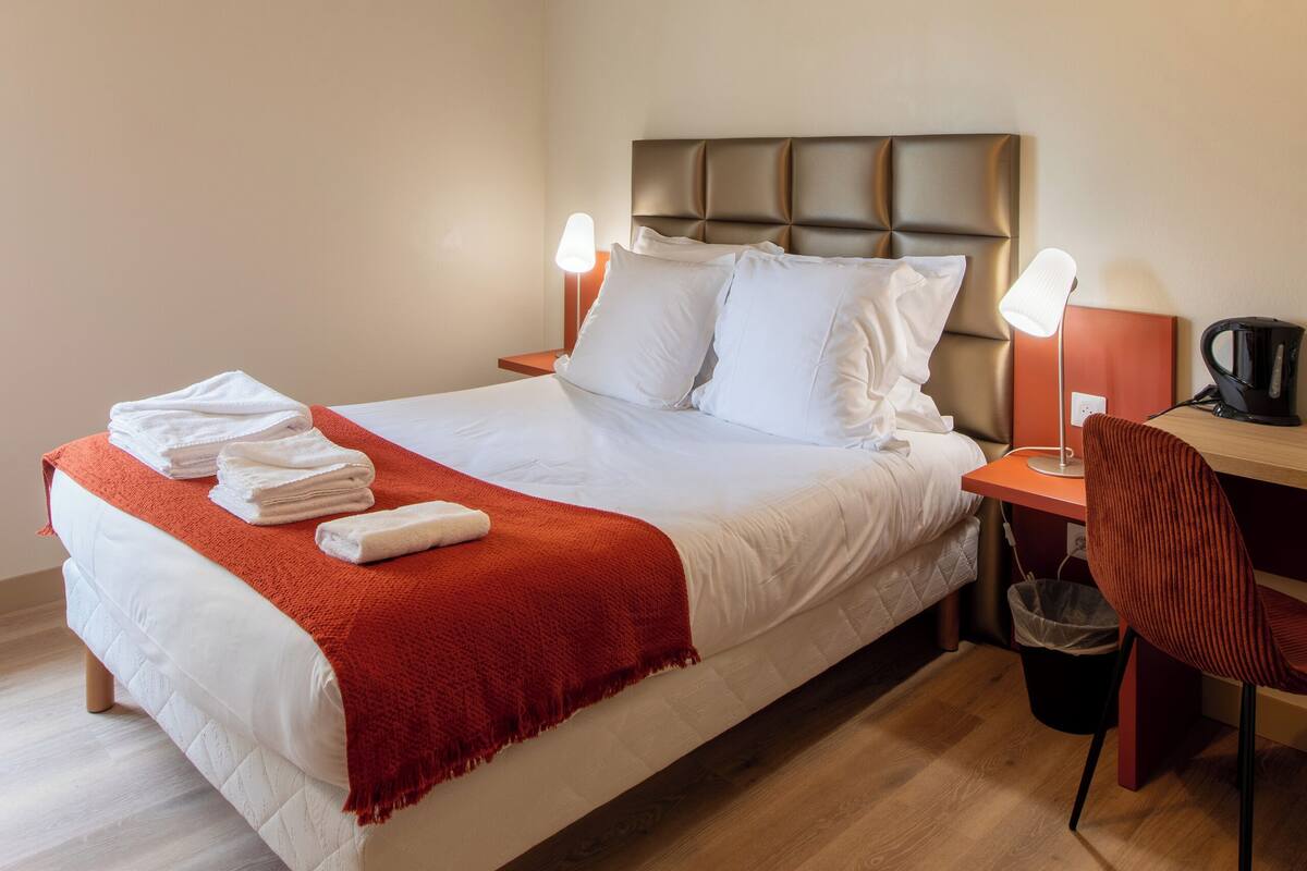 A neatly made bed with white linens and a red throw blanket, a wooden floor, a modern ceiling light, and a small desk with a chair.