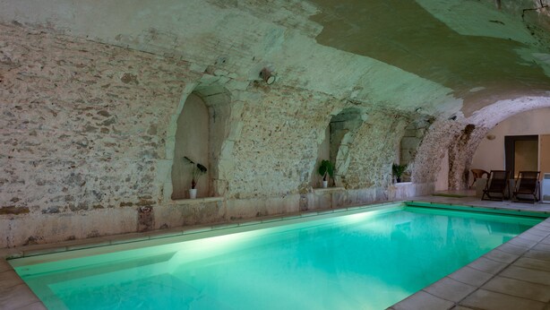 A swimming pool with stone walls and arches, potted plants, and a few chairs.