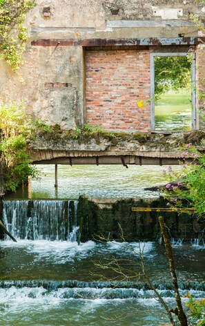 Un puente de piedra rústico sobre un río caudaloso, con una estructura de ladrillo a la derecha y vegetación circundante.