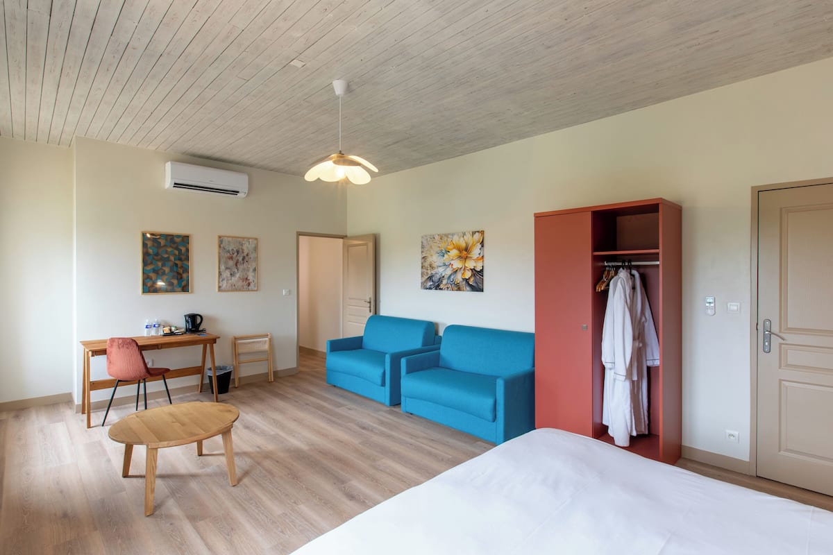 A room with a bed, two blue sofas, a wooden table, a desk with a chair, and a wardrobe.