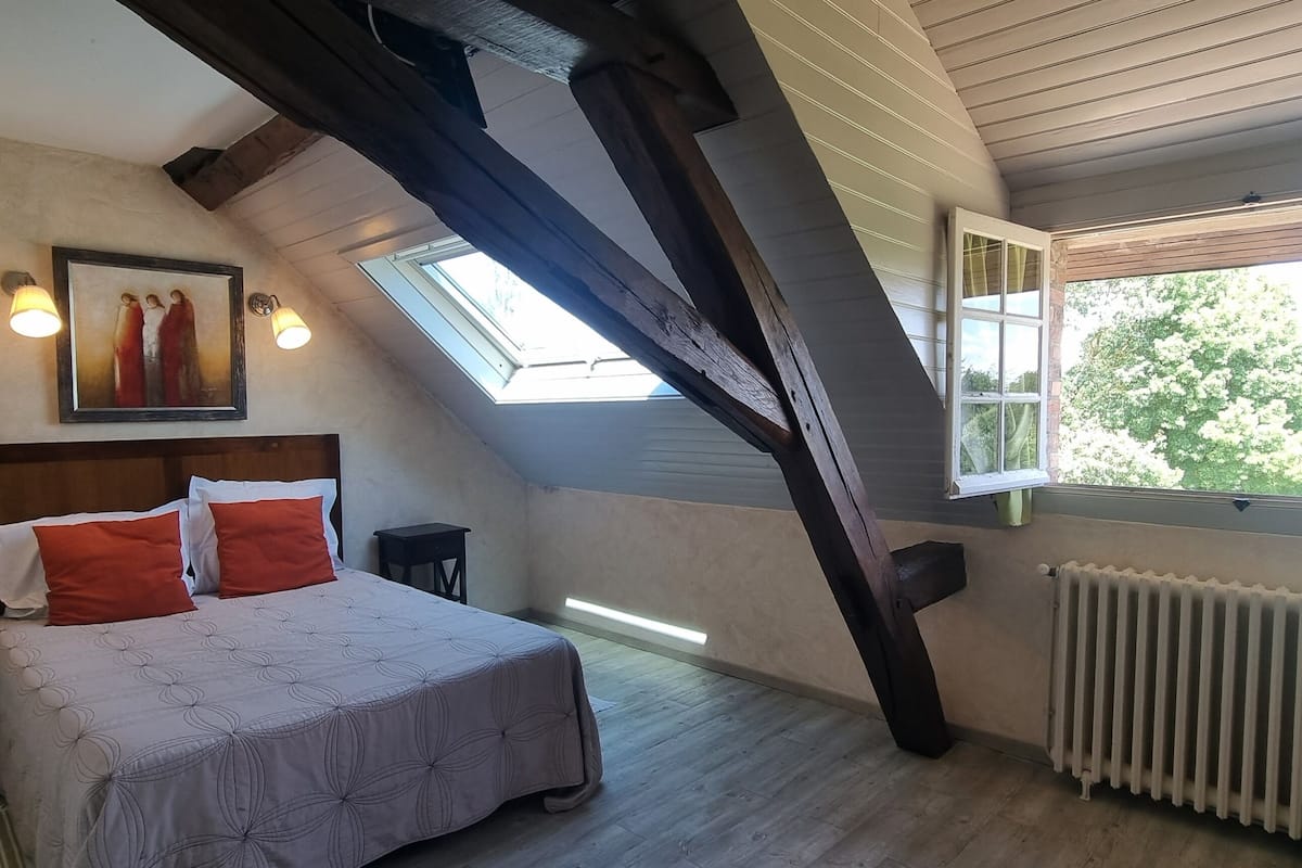 A bedroom with a sloped ceiling, a bed with red and white pillows, a nightstand, a framed picture on the wall, and a window with a view of trees.