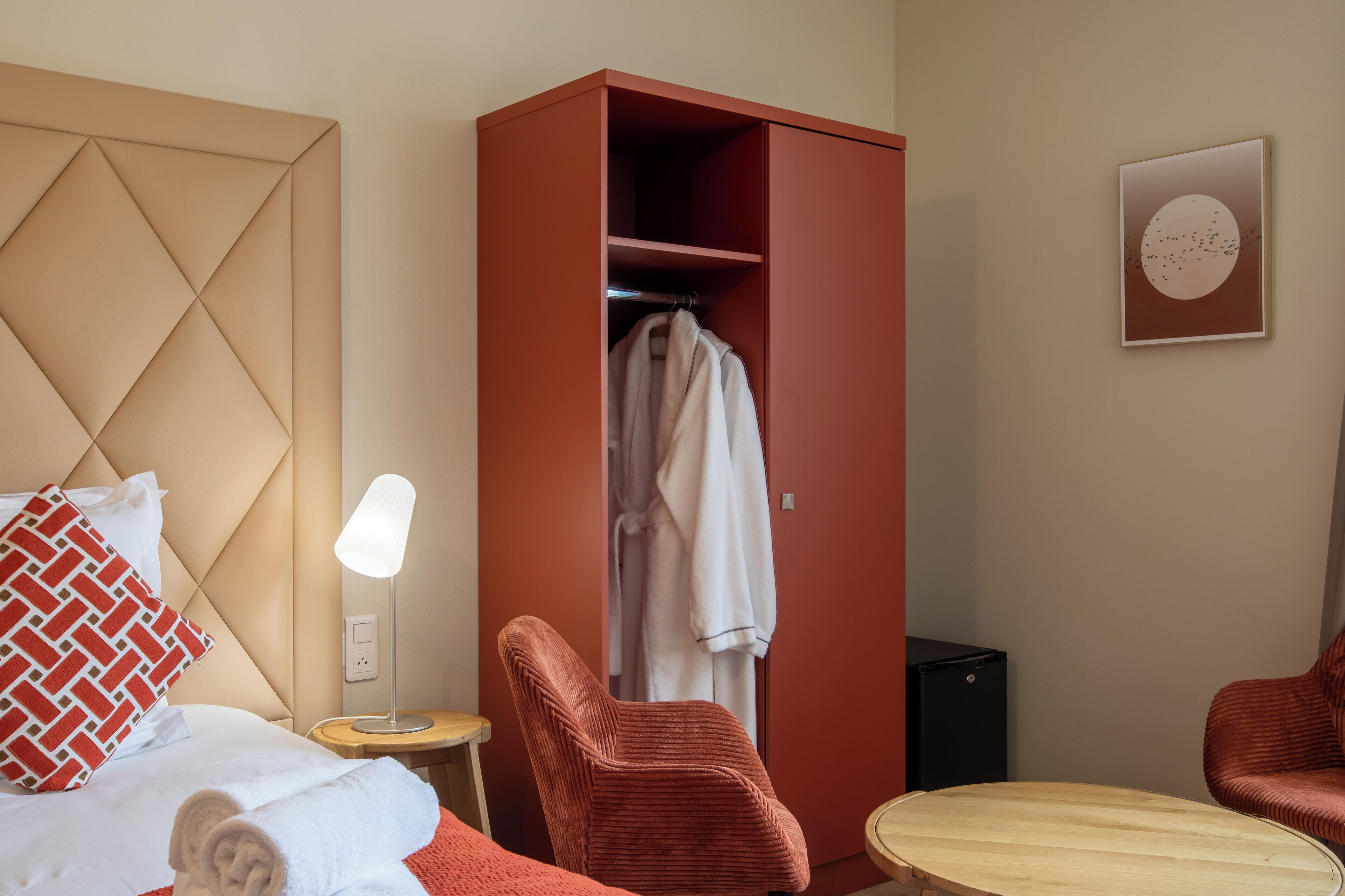 Hotellihuone, jossa on sänky, punainen vaatekaappi, kaksi punaista tuolia, pyöreä puinen pöytä, yöpöytälamppu ja seinälle ripustettu taulu.