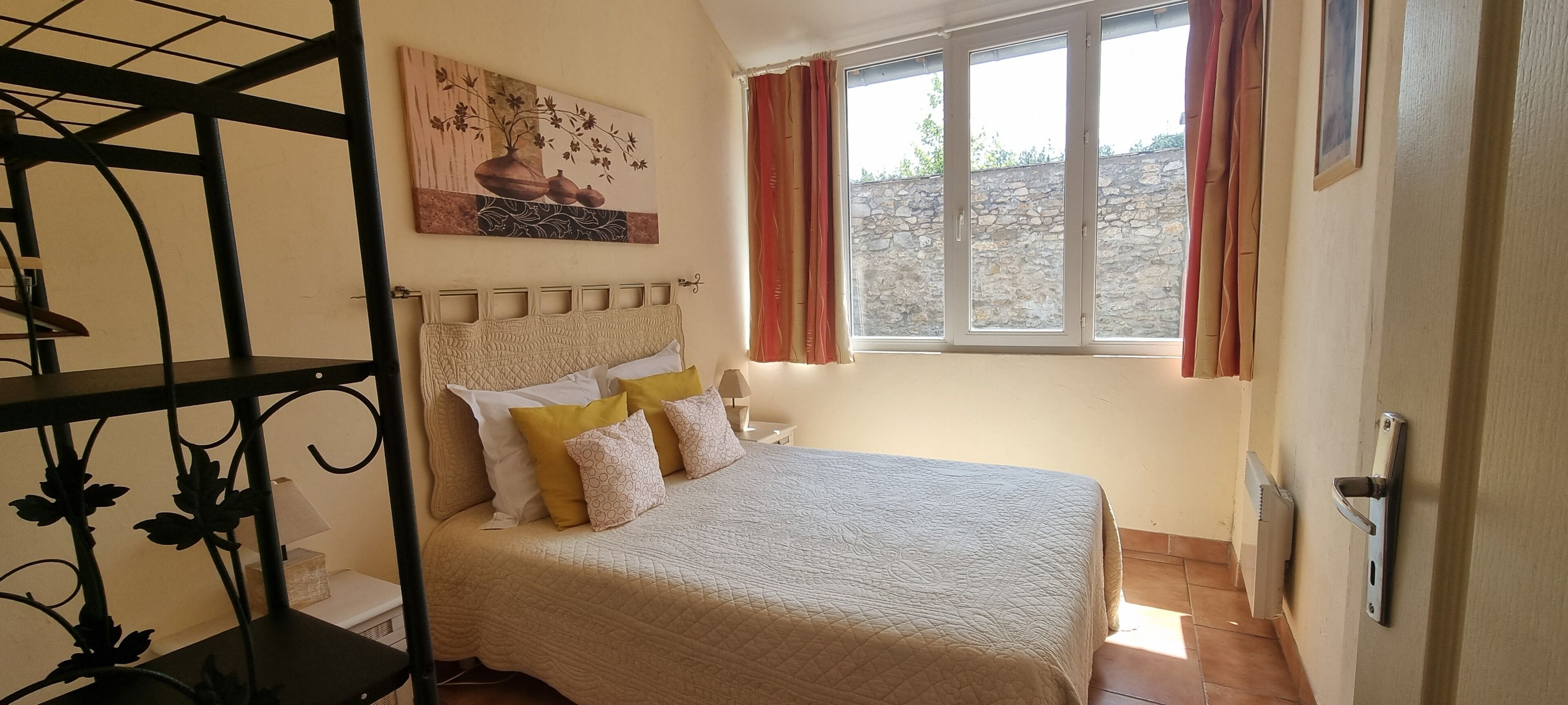 Un dormitorio con cama, cabecera, ventana con cortinas, un cuadro en la pared y una estantería de metal.