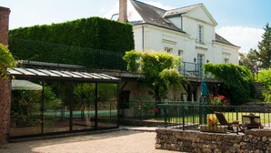 A covered outdoor area with a glass enclosure, a stone wall, and a white building with a green roof in the background.