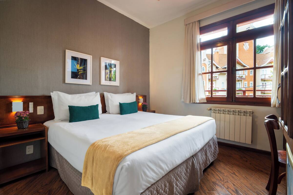 Een hotelkamer met een groot bed, twee nachtkastjes, een raam met uitzicht op gebouwen, en twee ingelijste foto's aan de muur.