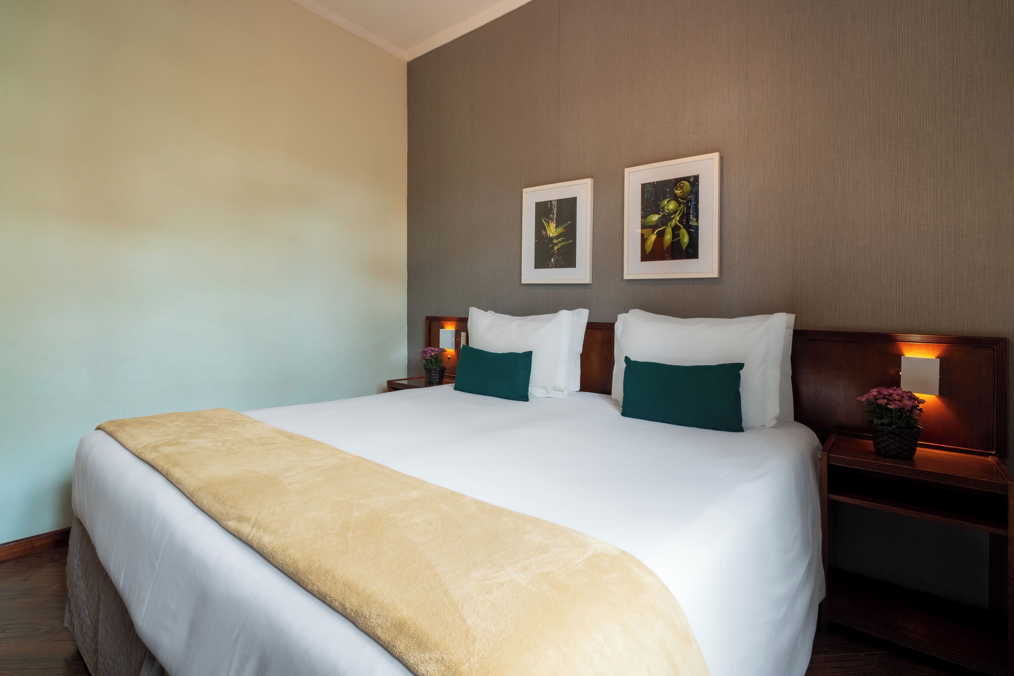 A neatly made bed with a white and beige blanket, two green pillows, and a wooden headboard. There are two framed pictures on the wall above the bed.