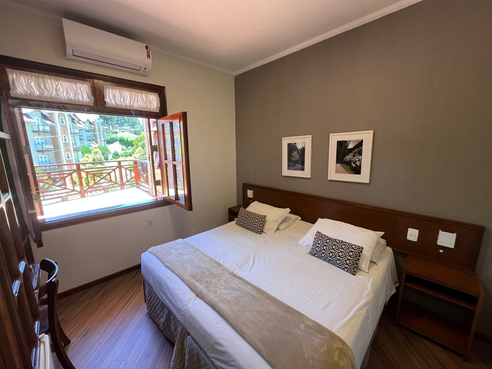 Un dormitorio con una cama grande, una ventana con vistas a edificios y vegetación, dos cuadros en la pared y un cabecero de madera.