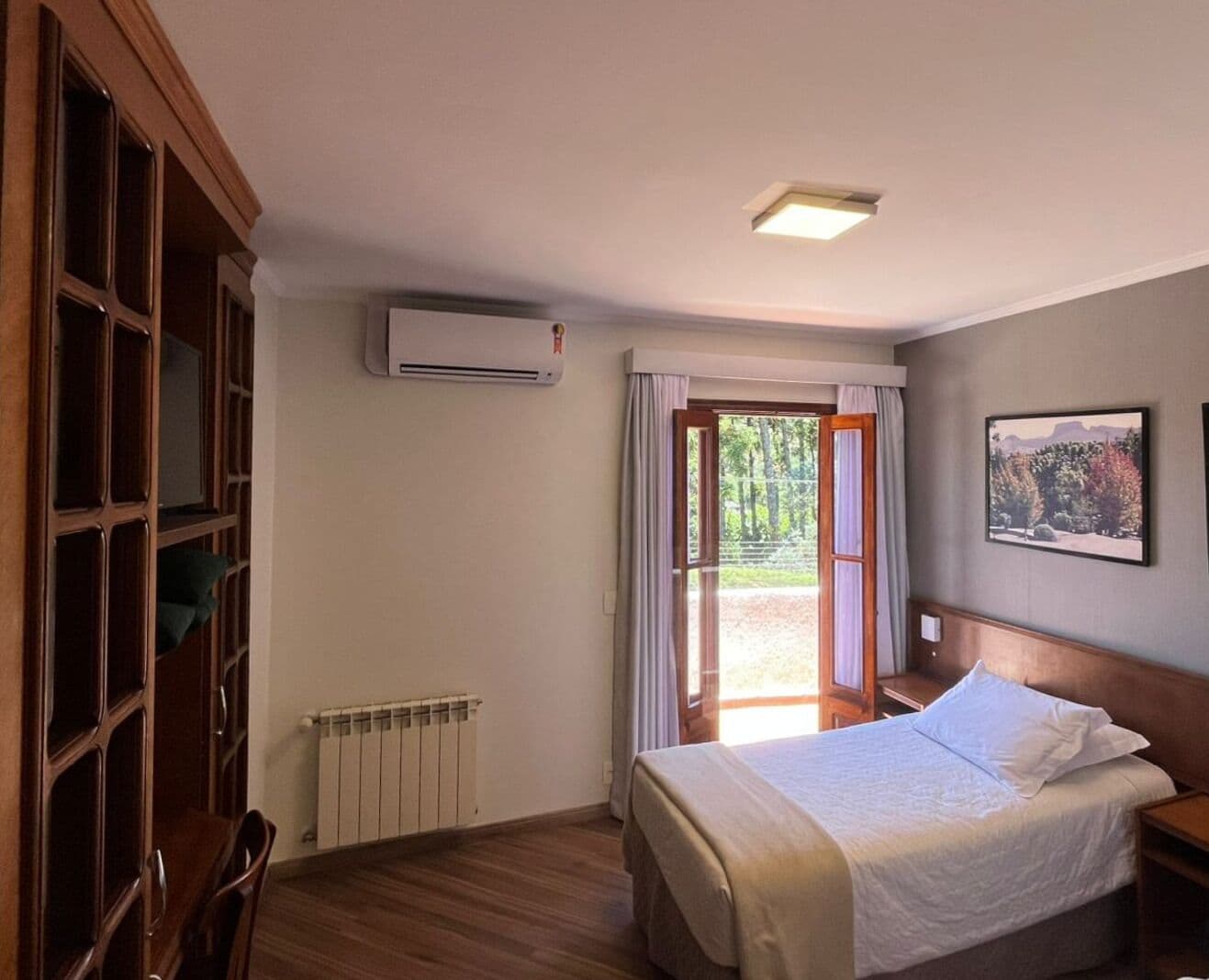 A hotel room with a bed, a radiator, an air conditioning unit, a window with curtains, and a view of the outdoors.