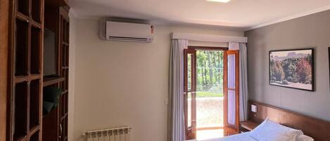 A hotel room with a bed, a radiator, an air conditioning unit, a window with curtains, and a view of the outdoors.