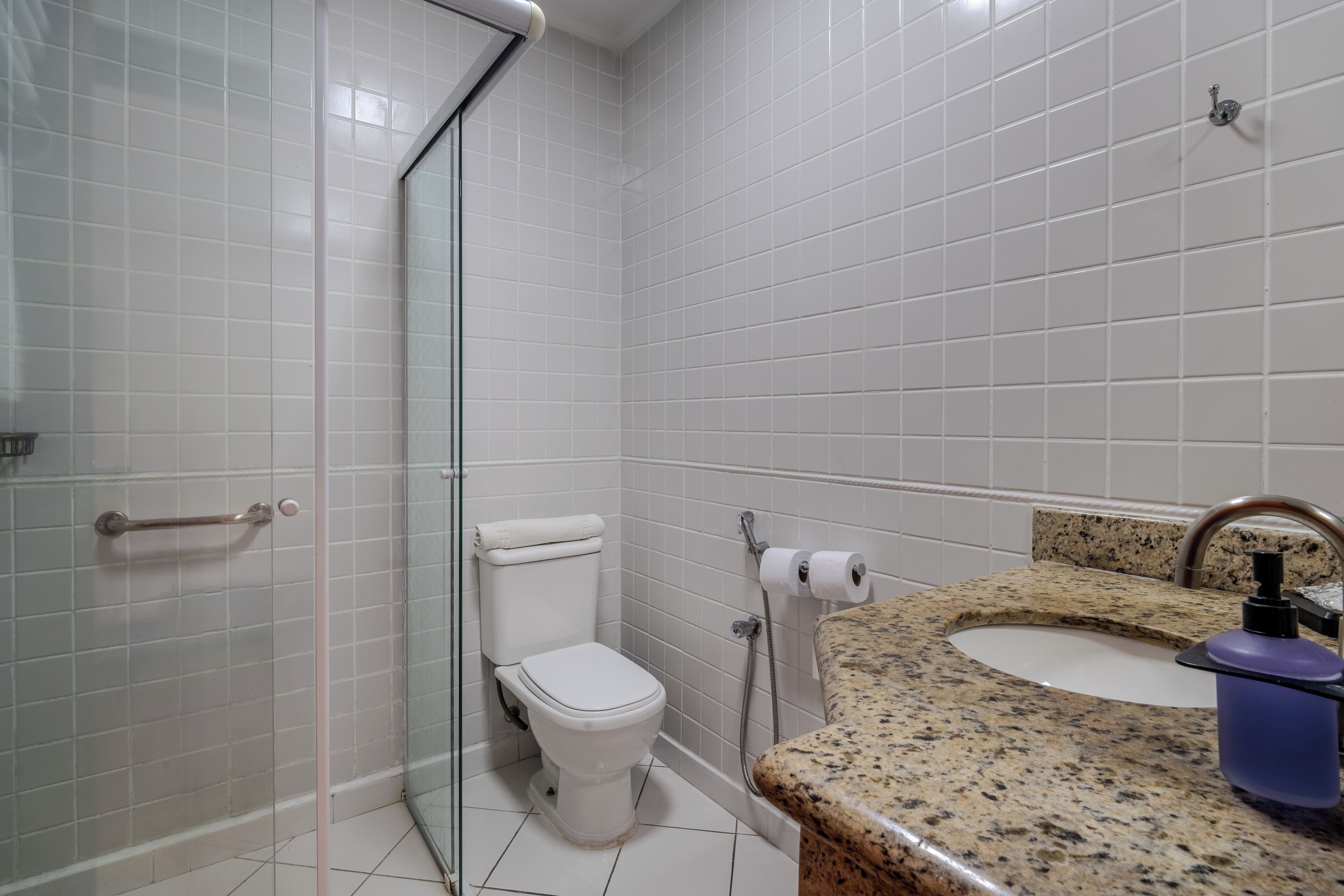 Un baño con cabina de ducha de vidrio, un inodoro blanco, encimera de granito con lavabo y dispensador de jabón.
