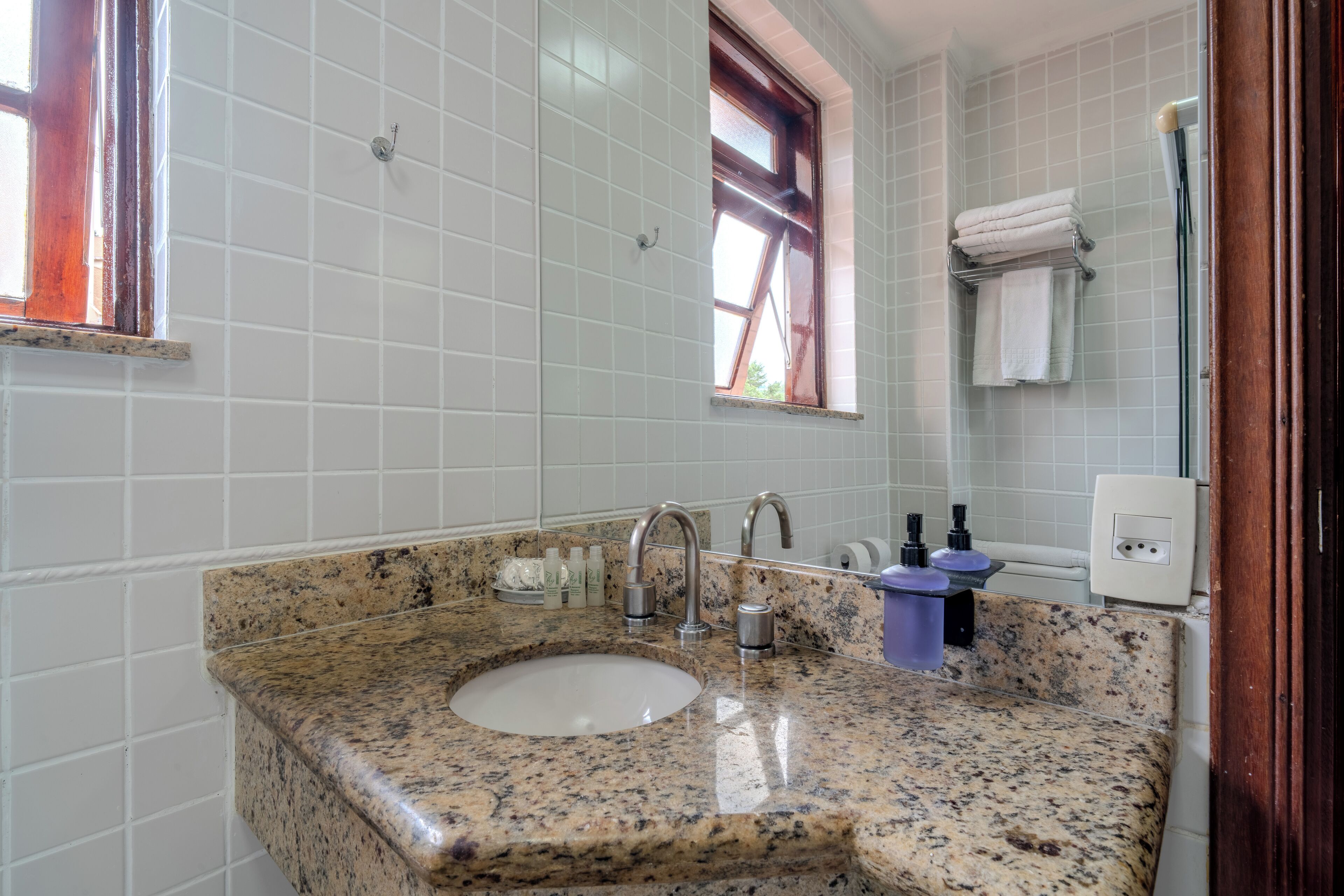 A bathroom with a granite sink, white tiled walls, a window, and a towel rack.