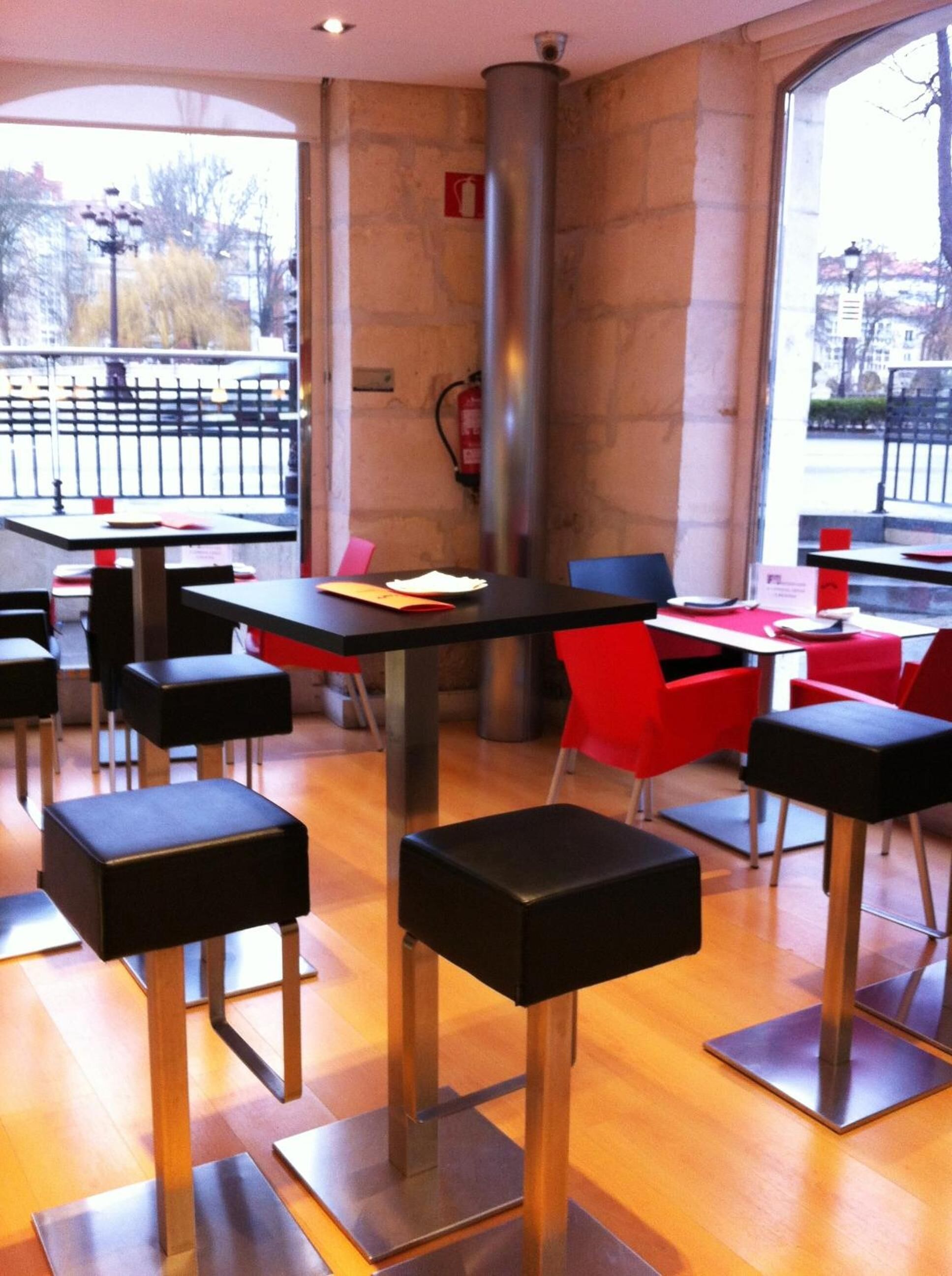 Un comedor moderno con sillas negras y rojas, mesas de madera y grandes ventanales que dan a la calle.
