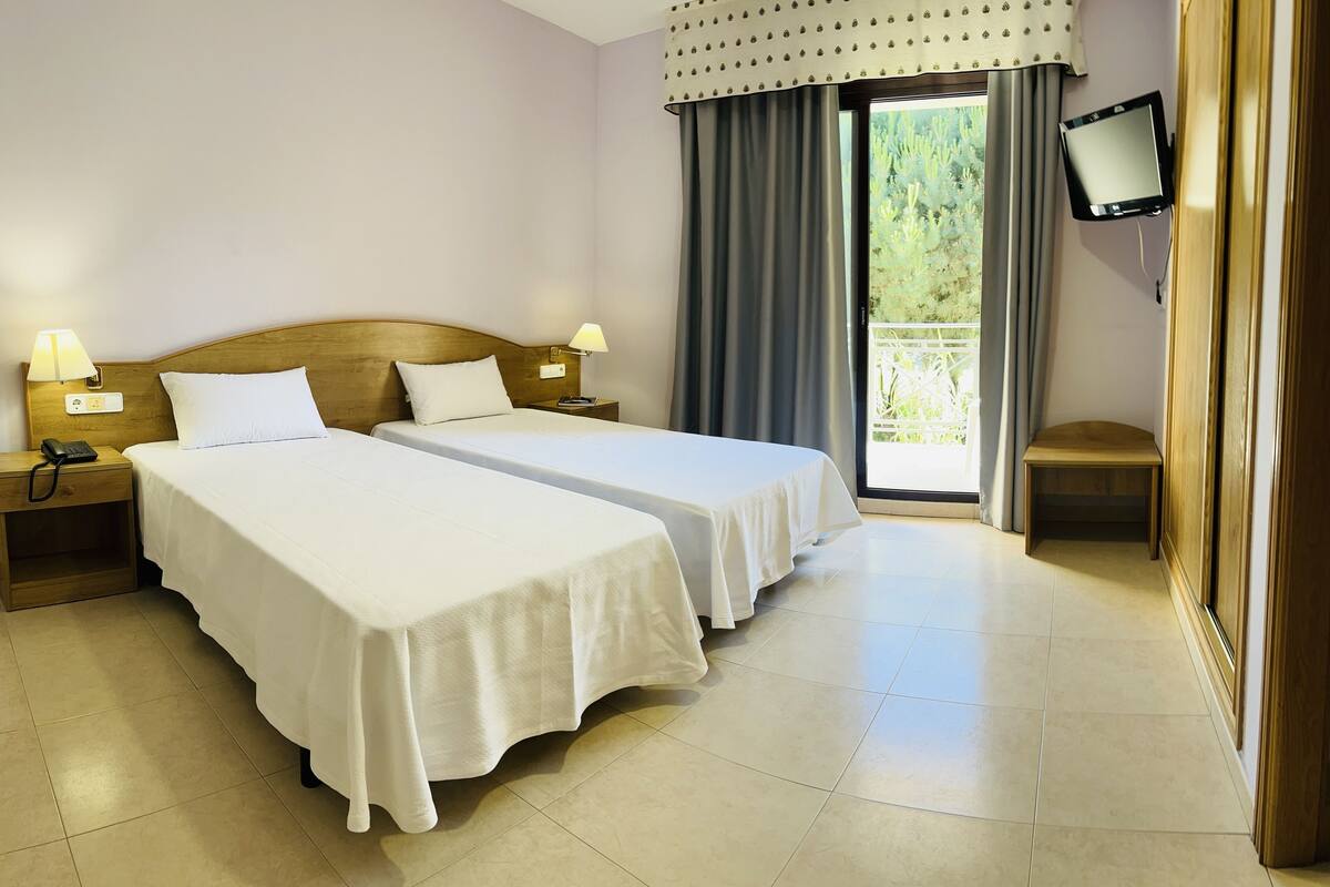 A hotel room with two beds, a bedside table with a lamp, a bedside phone, a TV on the wall, and a view of greenery through the window.