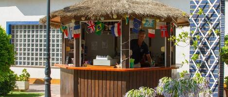 A small outdoor kiosk with a thatched roof, surrounded by potted plants and flags.