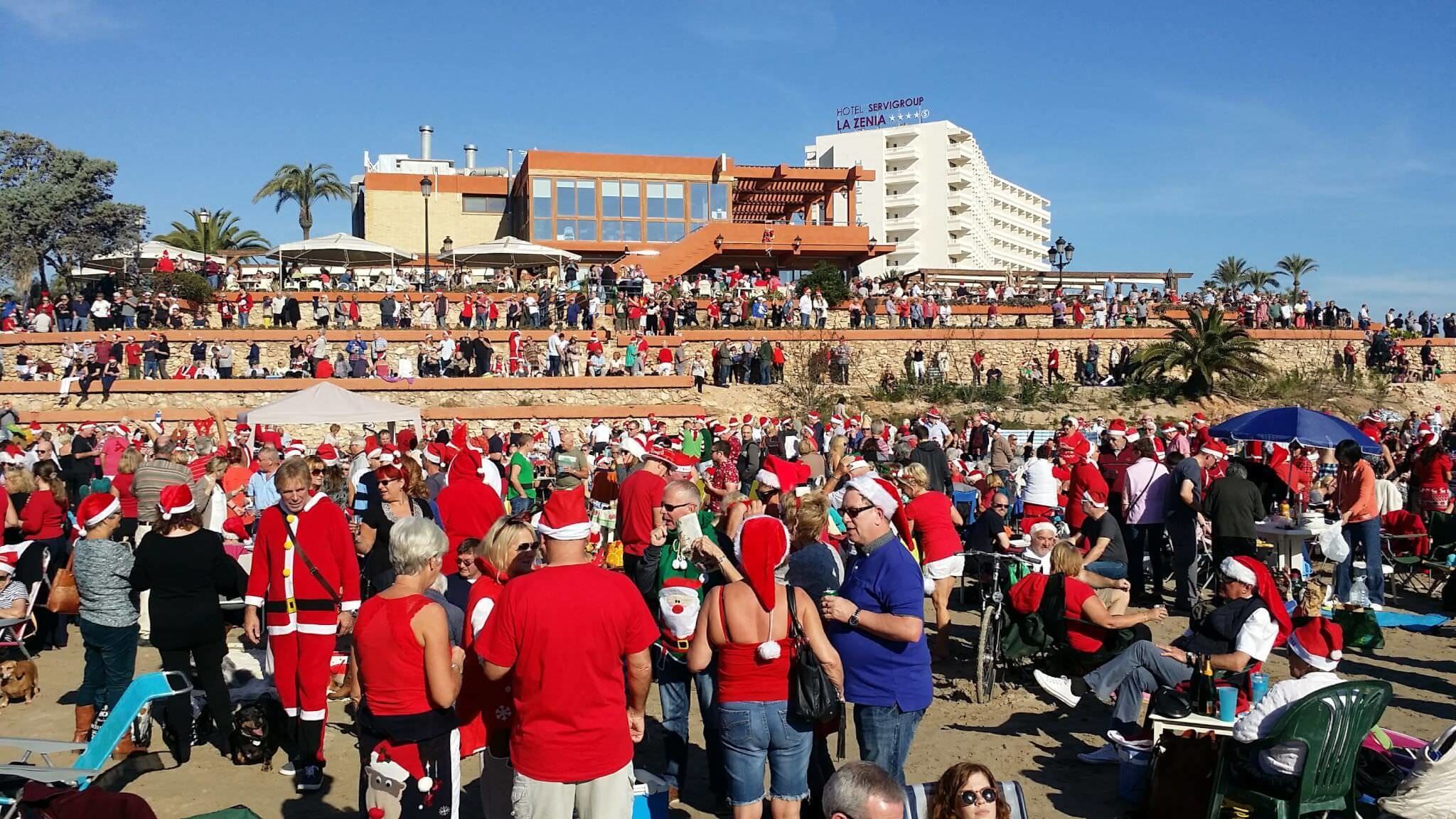 Evento al aire libre concurrido donde la gente viste ropa festiva, con el hotel 'Hotel Servigroup La Zenia' al fondo, y un cielo despejado.