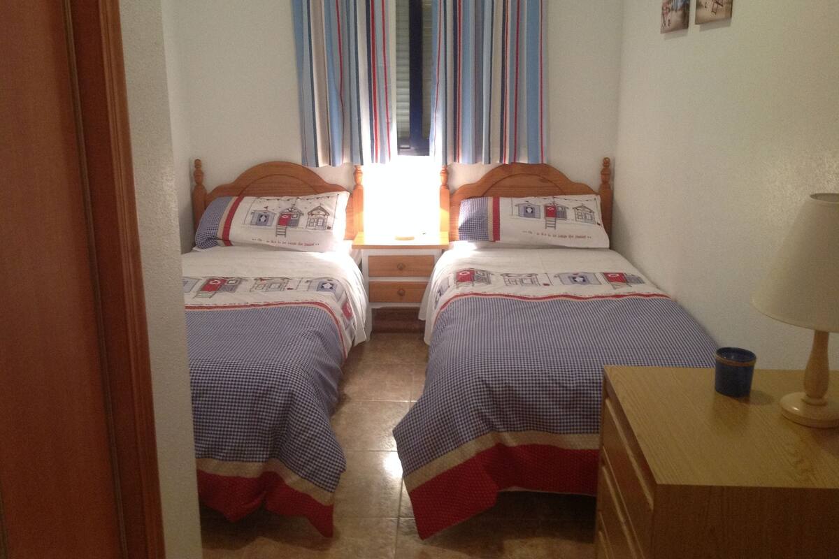 Un dormitorio con dos camas individuales, una mesita de noche de madera, una lámpara y una ventana con cortinas a rayas.
